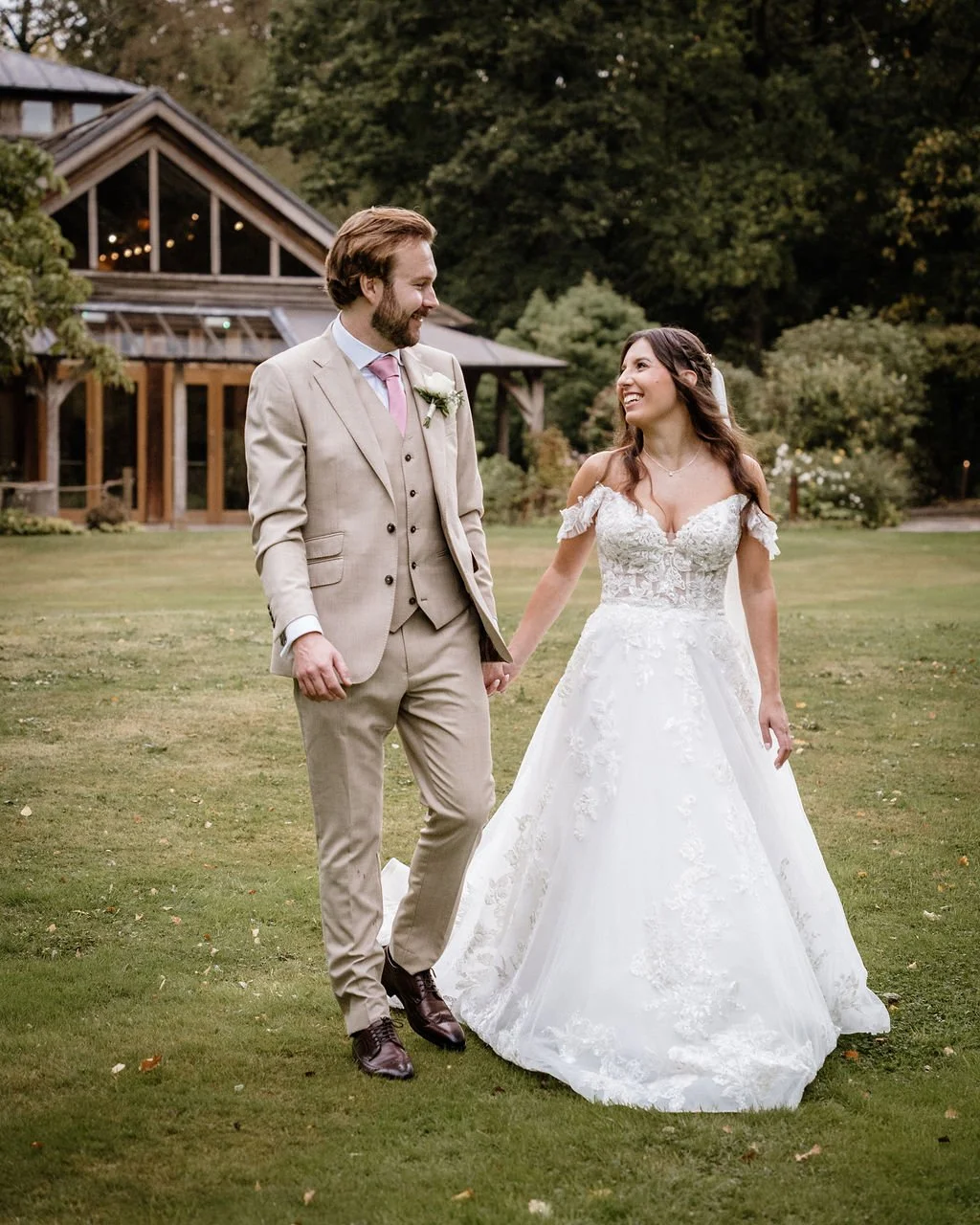 A bride and groom walking hand-in-hand on a grassy lawn in a scenic outdoor setting with a house and trees in the background, both smiling at each other.