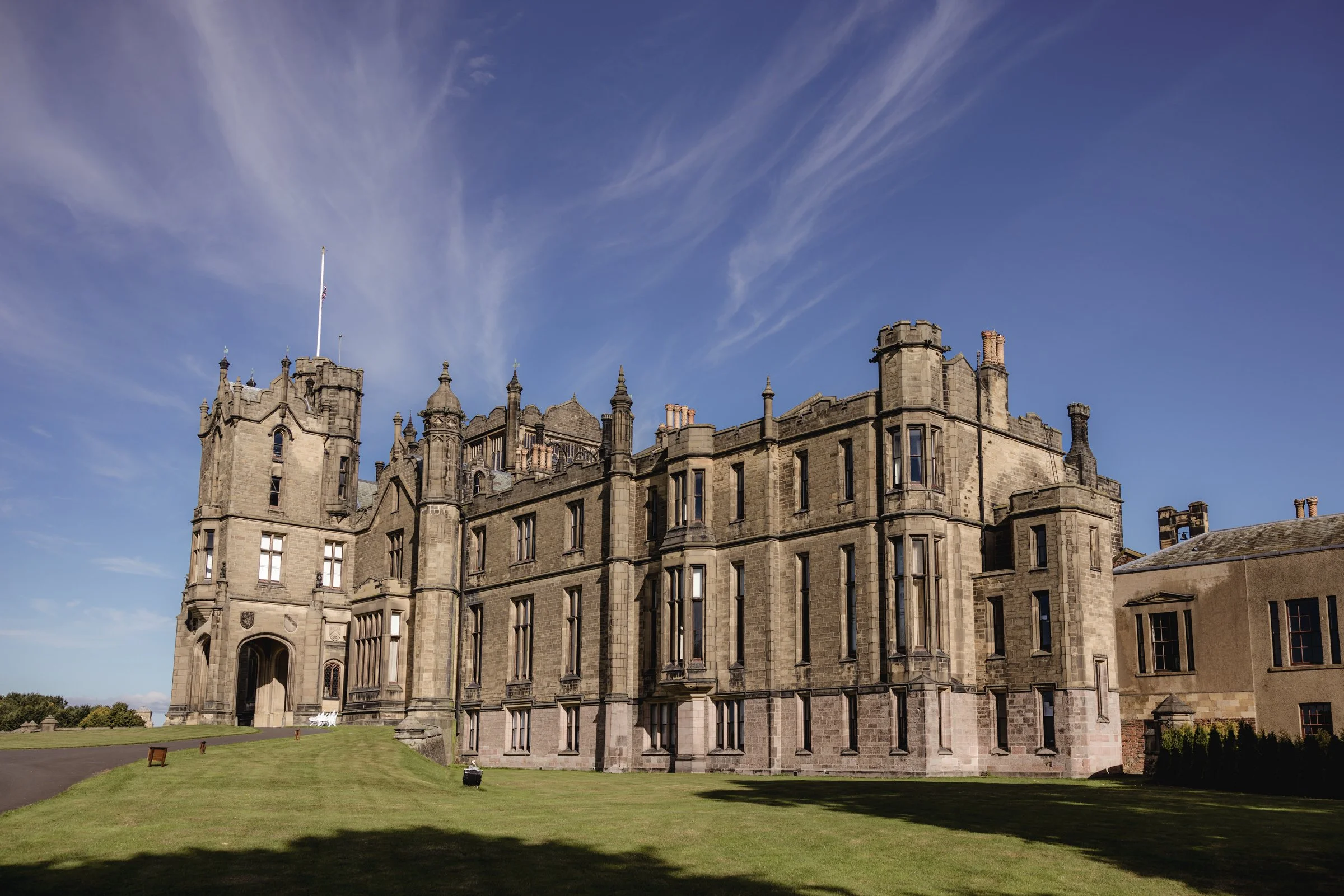 A large historic castle made of gray stone, with multiple towers and turrets, situated on a lush green lawn under a partly cloudy blue sky.