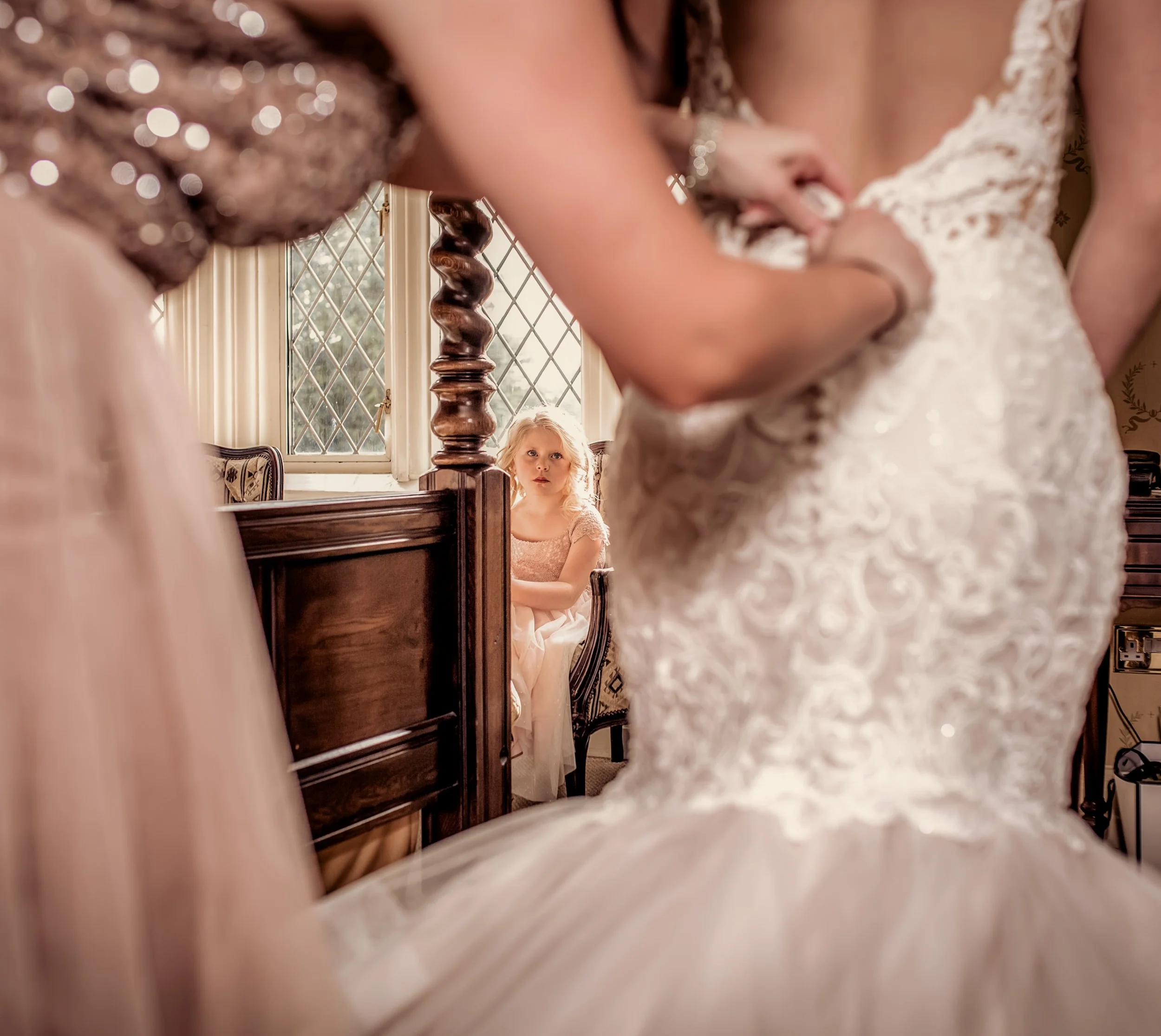 A young girl with blonde hair wearing a pink dress, sitting on a chair and watching a wedding ceremony through a gap between two women in wedding dresses inside a room with wooden furniture and diamond-patterned window panes.