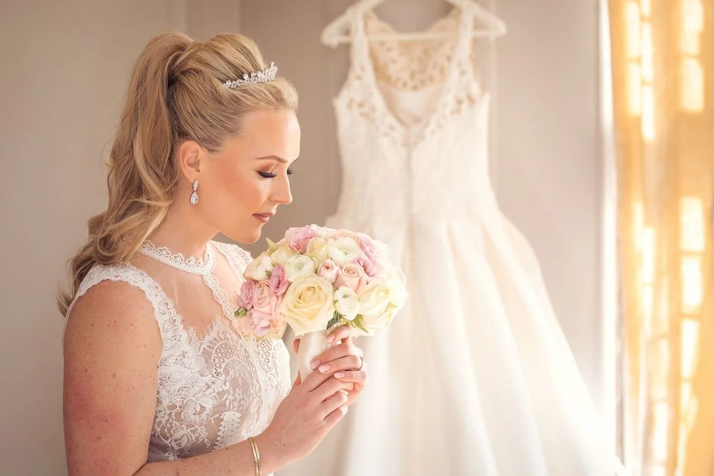 Bride in lace dress holding a bouquet of pink and white roses, standing in front of a hanging wedding dress, near a window with yellow curtains.