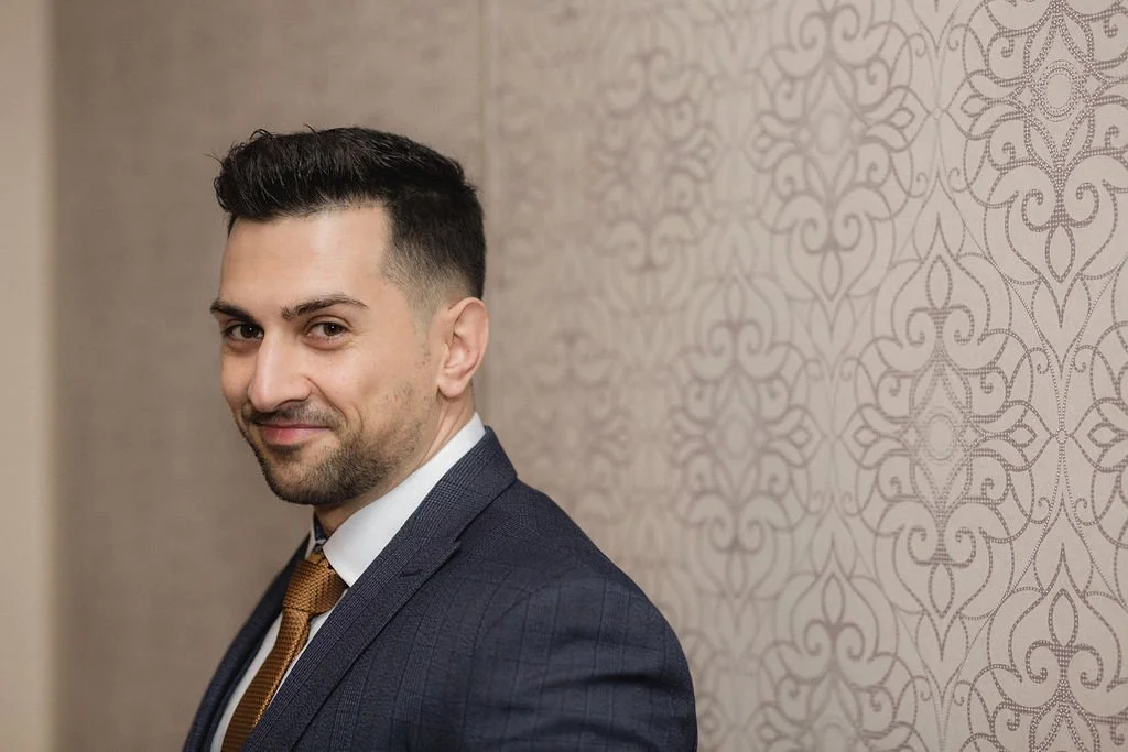 A man in a dark suit with a white shirt and gold tie, standing against a patterned wall, smiling slightly at the camera.