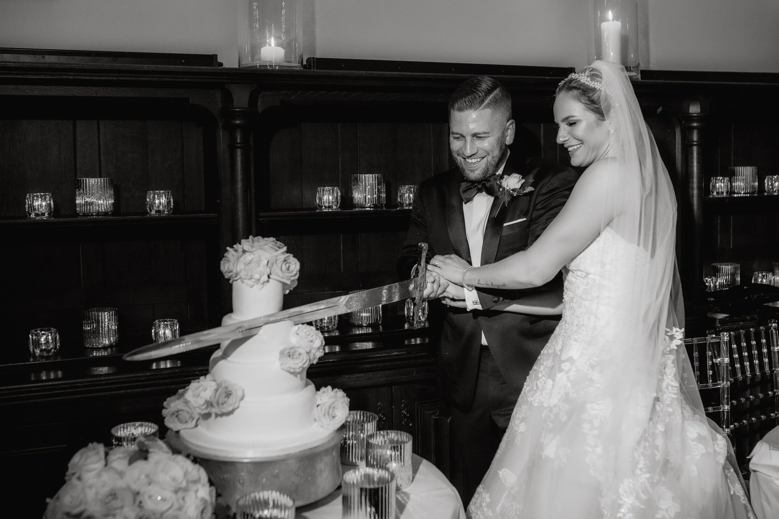 A newlywed couple cutting a wedding cake with a cake knife at their wedding reception, with candles and floral decorations in the background.