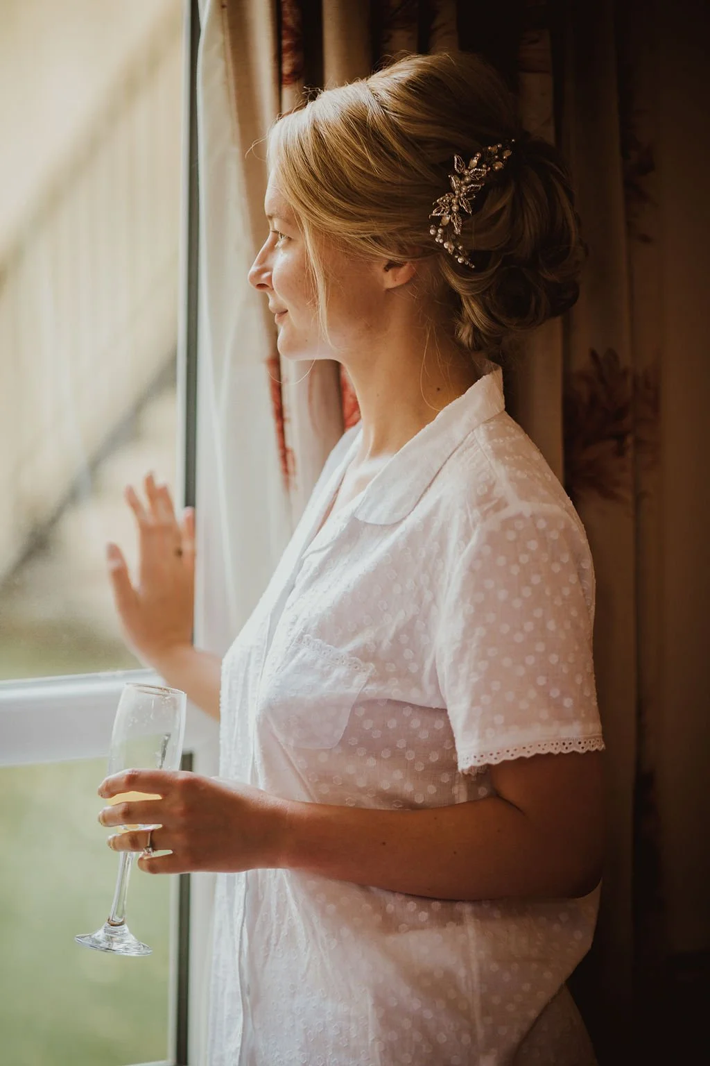 A woman with blonde hair in an elegant updo, wearing a decorative hair accessory, stands by a window holding a glass of champagne, looking outside on a cloudy day.