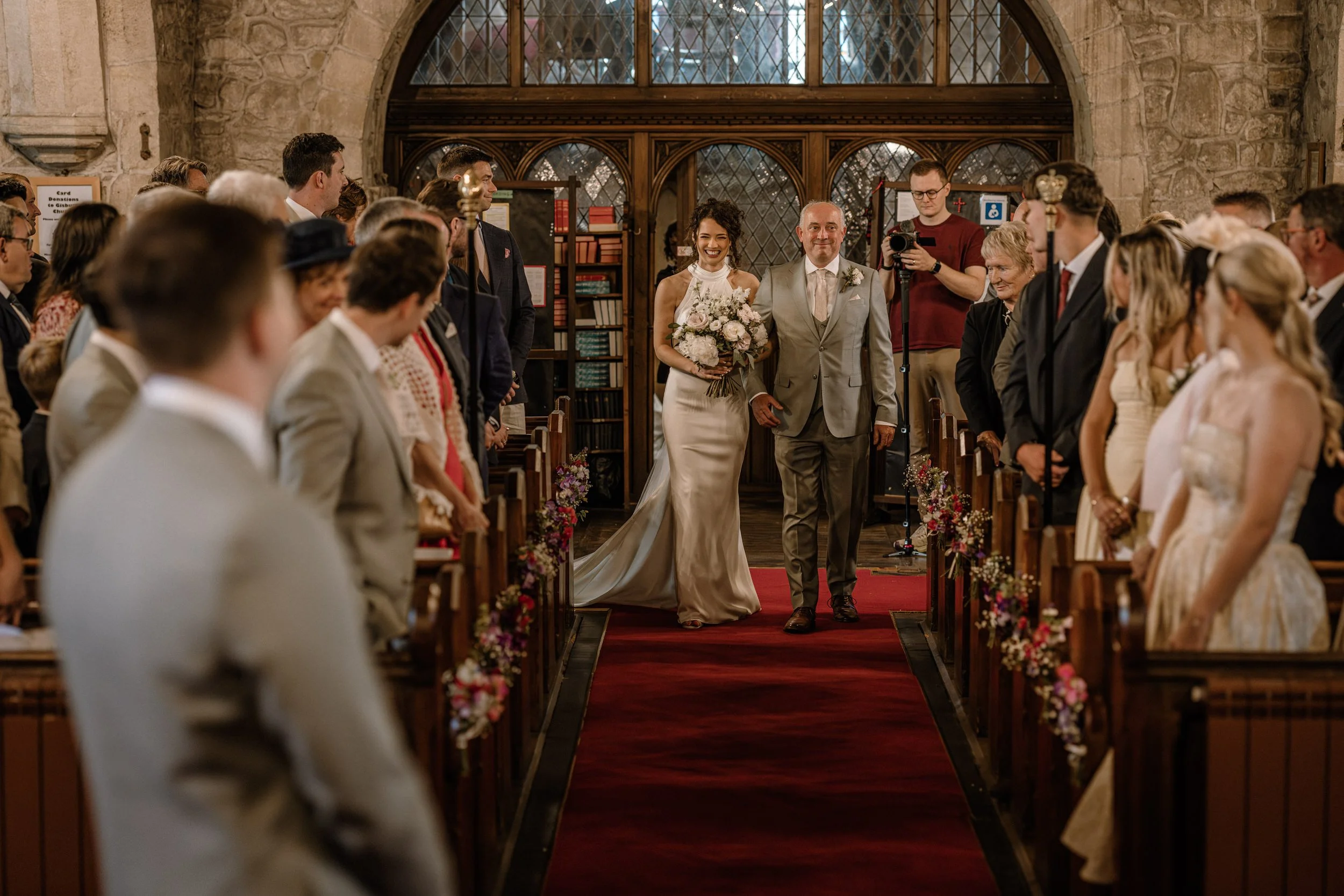 A bride and her father walking down the aisle in a church during a wedding ceremony, surrounded by guests seated on either side, with a photographer capturing the moment.