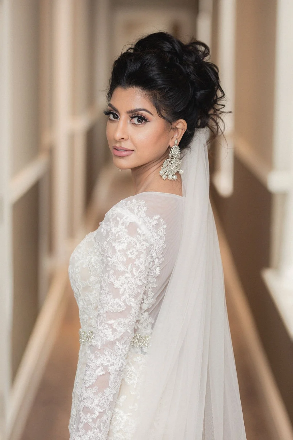 A woman in a white lace wedding dress with long sleeves, large earrings, and a veil, standing in a hallway with beige walls.