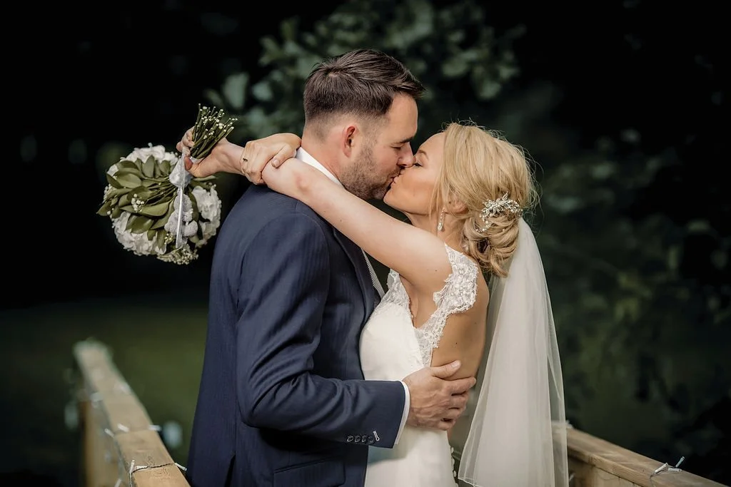A bride and groom share a kiss outdoors on a wooden bridge, with the bride holding a bouquet of flowers and wearing a lace wedding dress and veil, in front of a blurred green background.