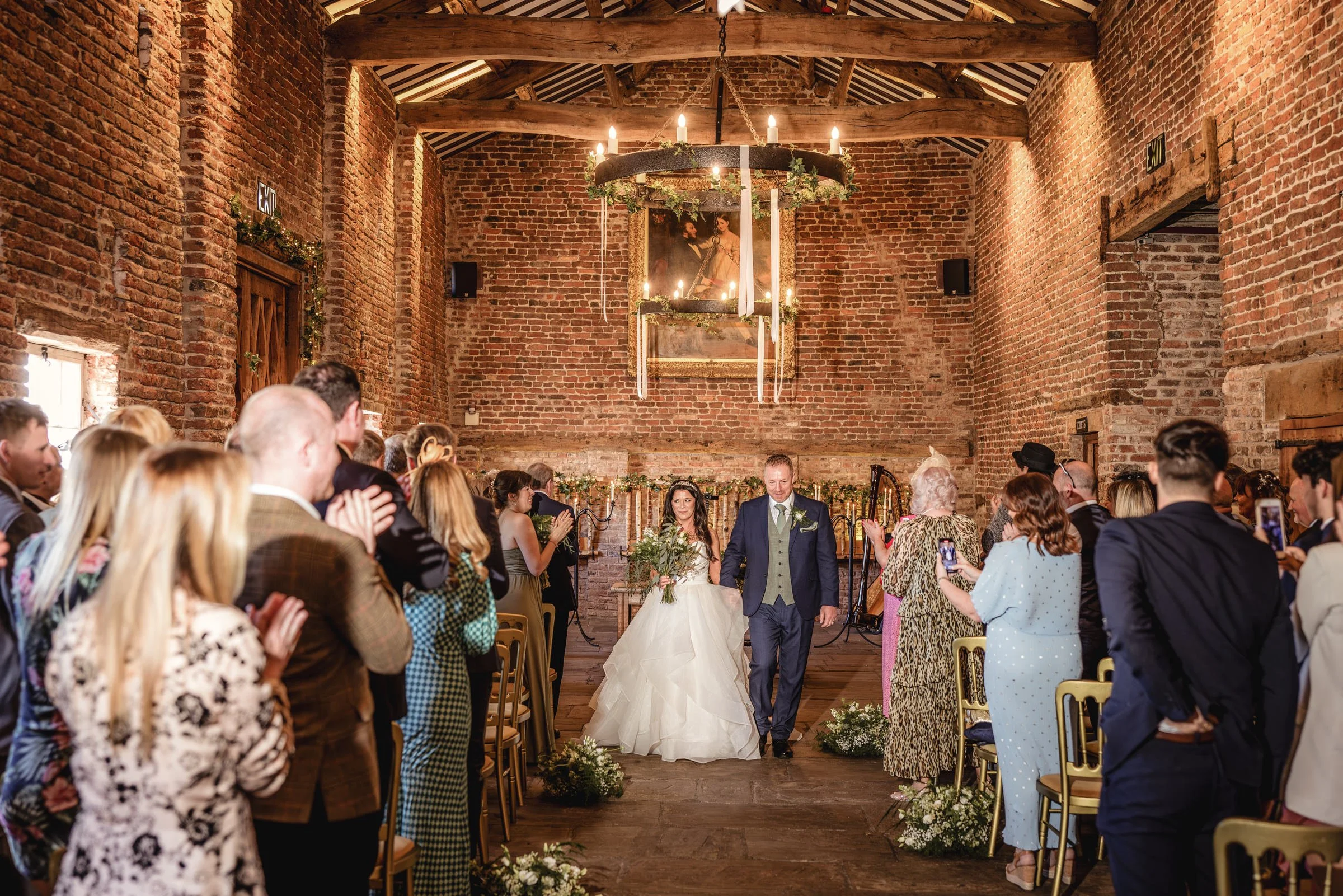 A bride and groom walking together down the aisle at their wedding ceremony inside a rustic brick-chambered venue, with guests standing and clapping on either side.