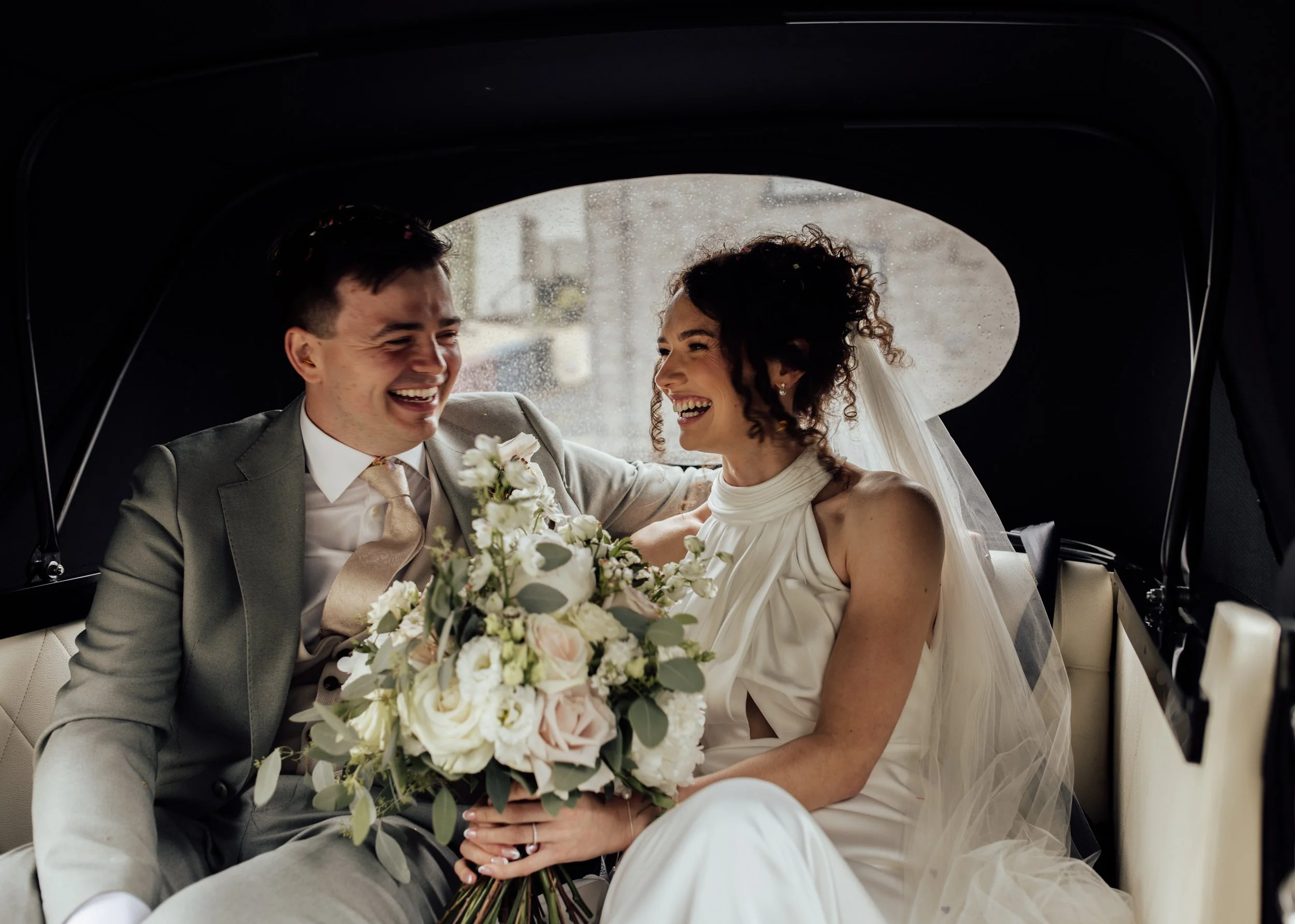 A smiling bride and groom sitting in the back of a car, the bride holding a bouquet of white and pink flowers, both dressed in wedding attire.