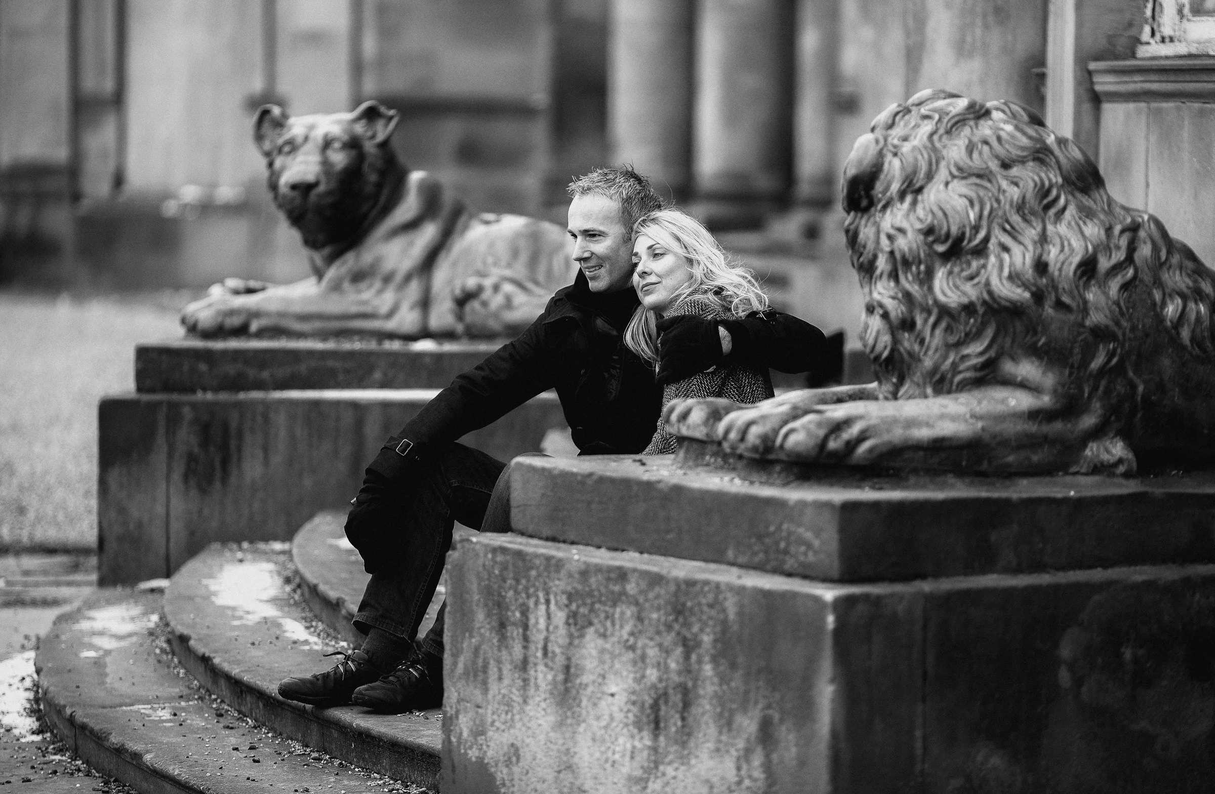 A black and white photo of a couple sitting on a stone step, leaning against a decorative stone ledge, surrounded by large stone lion sculptures in a park or garden setting.