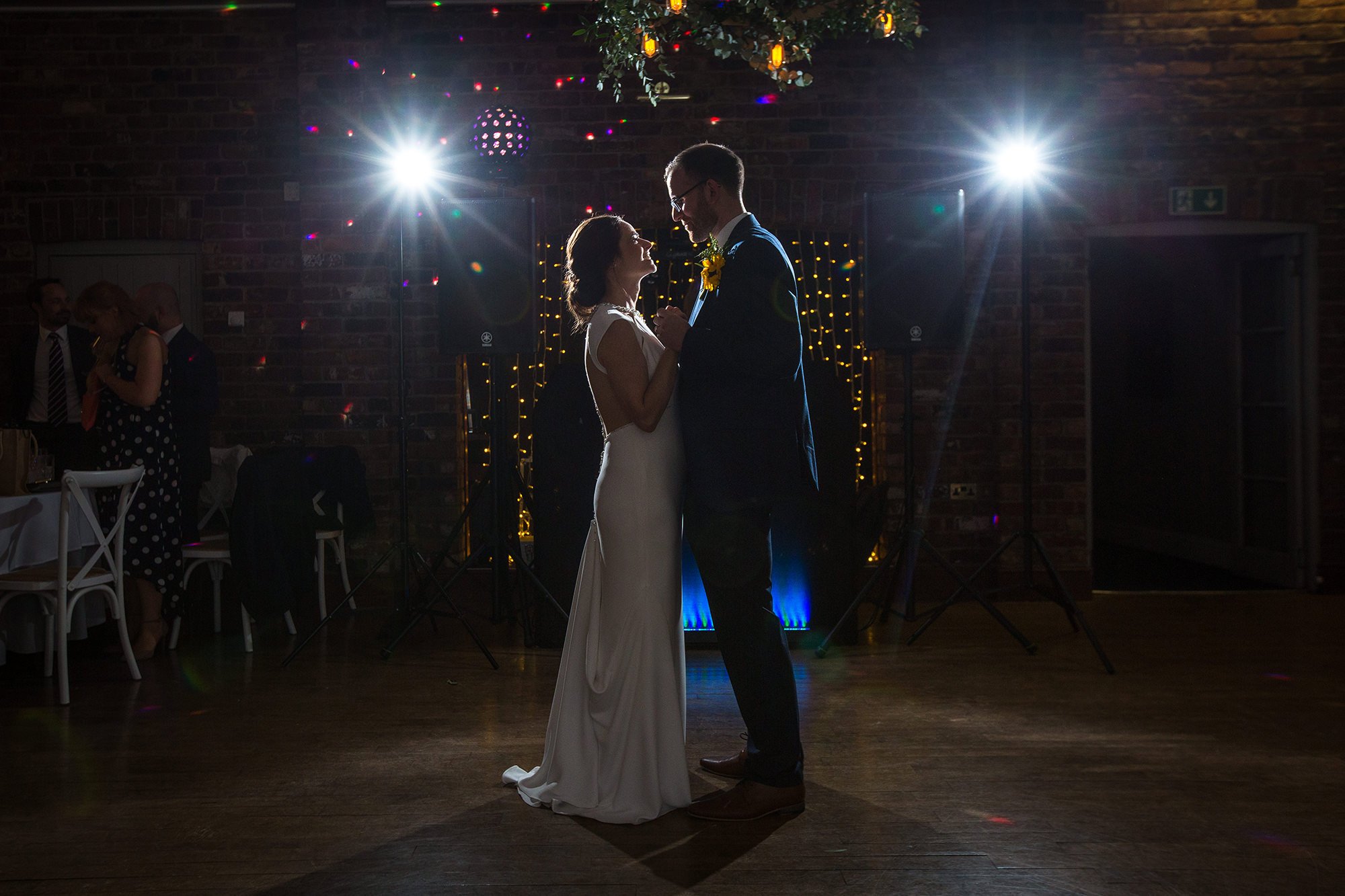 A bride and groom are dancing closely during their wedding reception, with bright backlighting and decorative lights in the background.