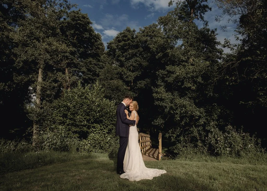 A couple in wedding attire embracing outdoors on a grassy area with dense trees in the background.