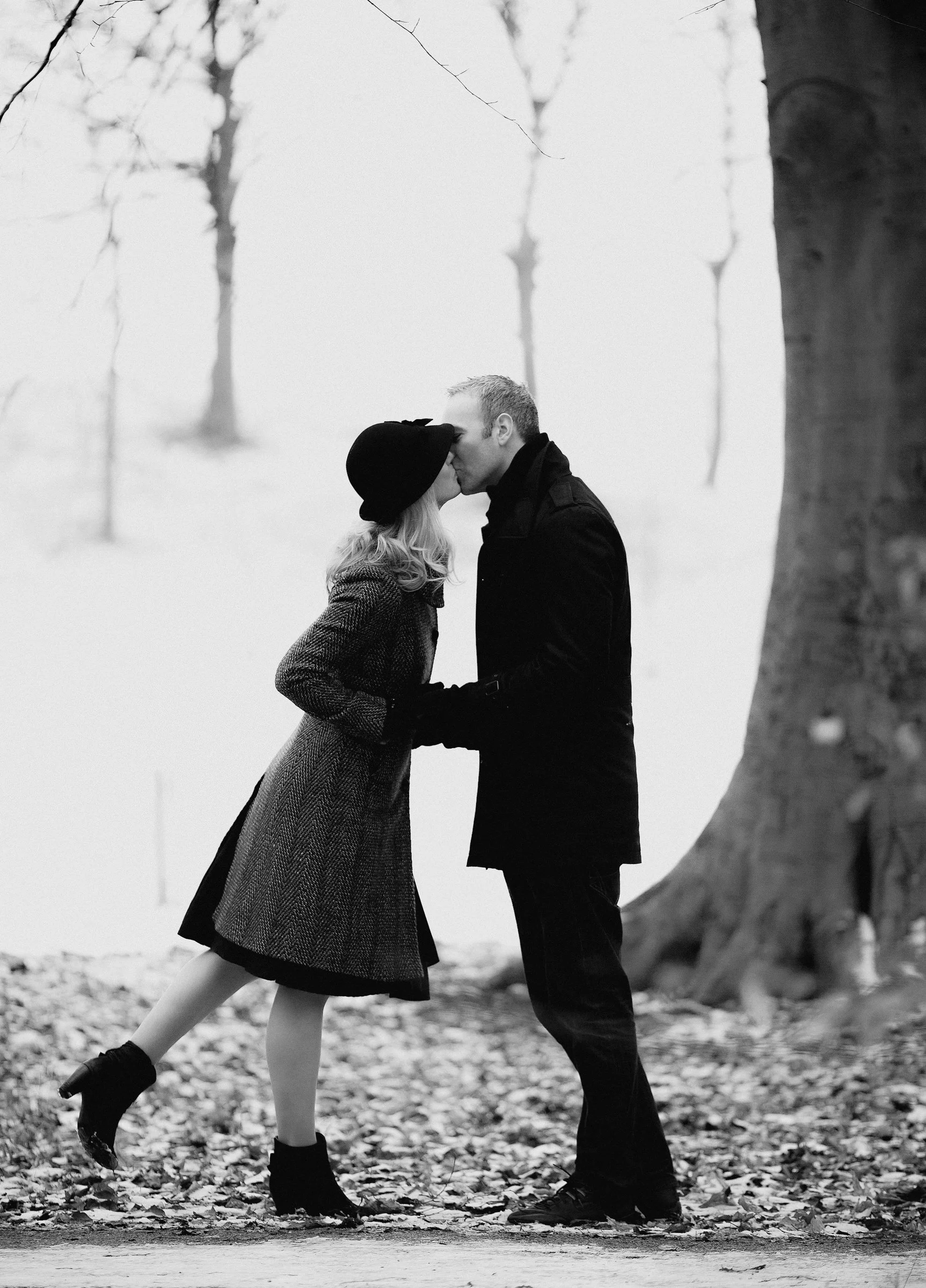 A black-and-white photo of a couple standing outdoors in a snowy landscape, sharing a kiss, with trees in the background and fallen leaves on the ground.