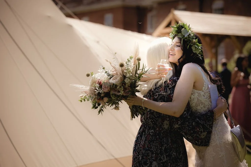 Two women hugging, one in a wedding dress wearing a floral crown, holding a champagne glass, and the other in a dark floral dress holding a bouquet of flowers, outdoors during a celebration.