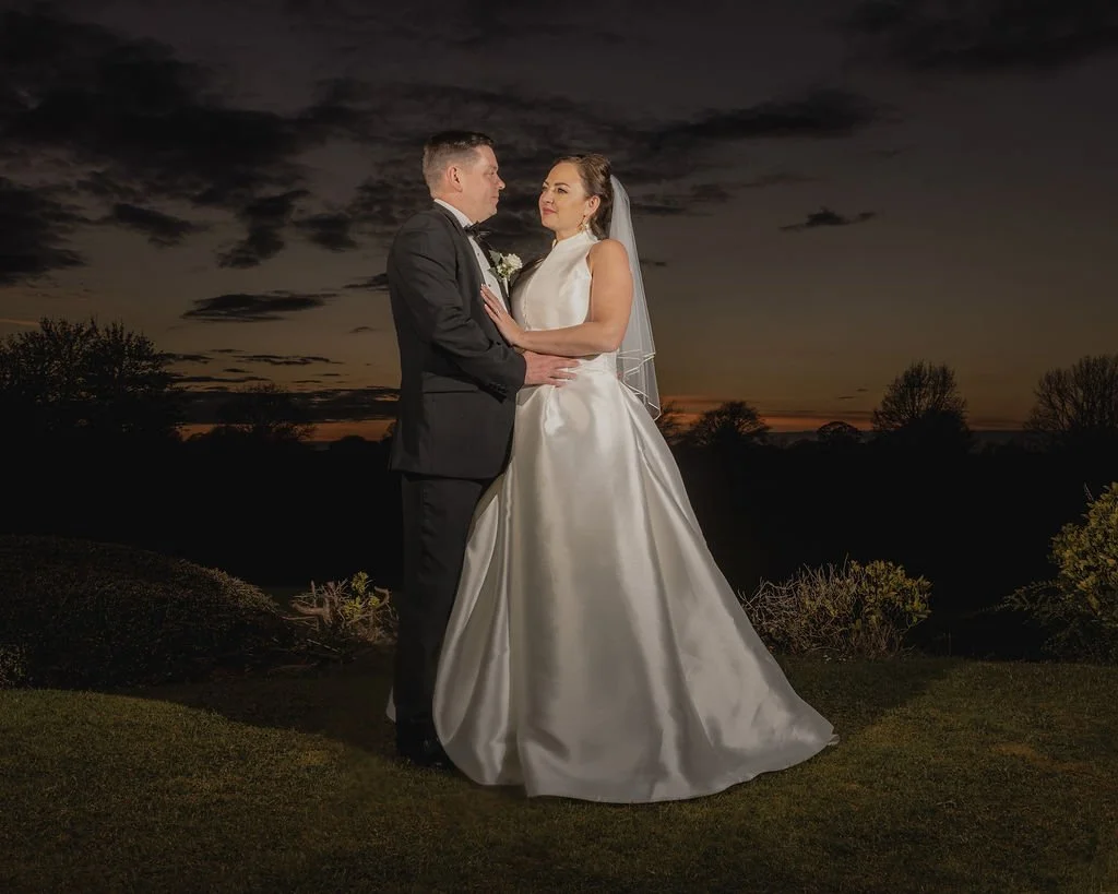 A bride and groom stand close together outdoors at sunset, with the bride wearing a white wedding gown and veil, and the groom in a black suit, facing each other lovingly.
