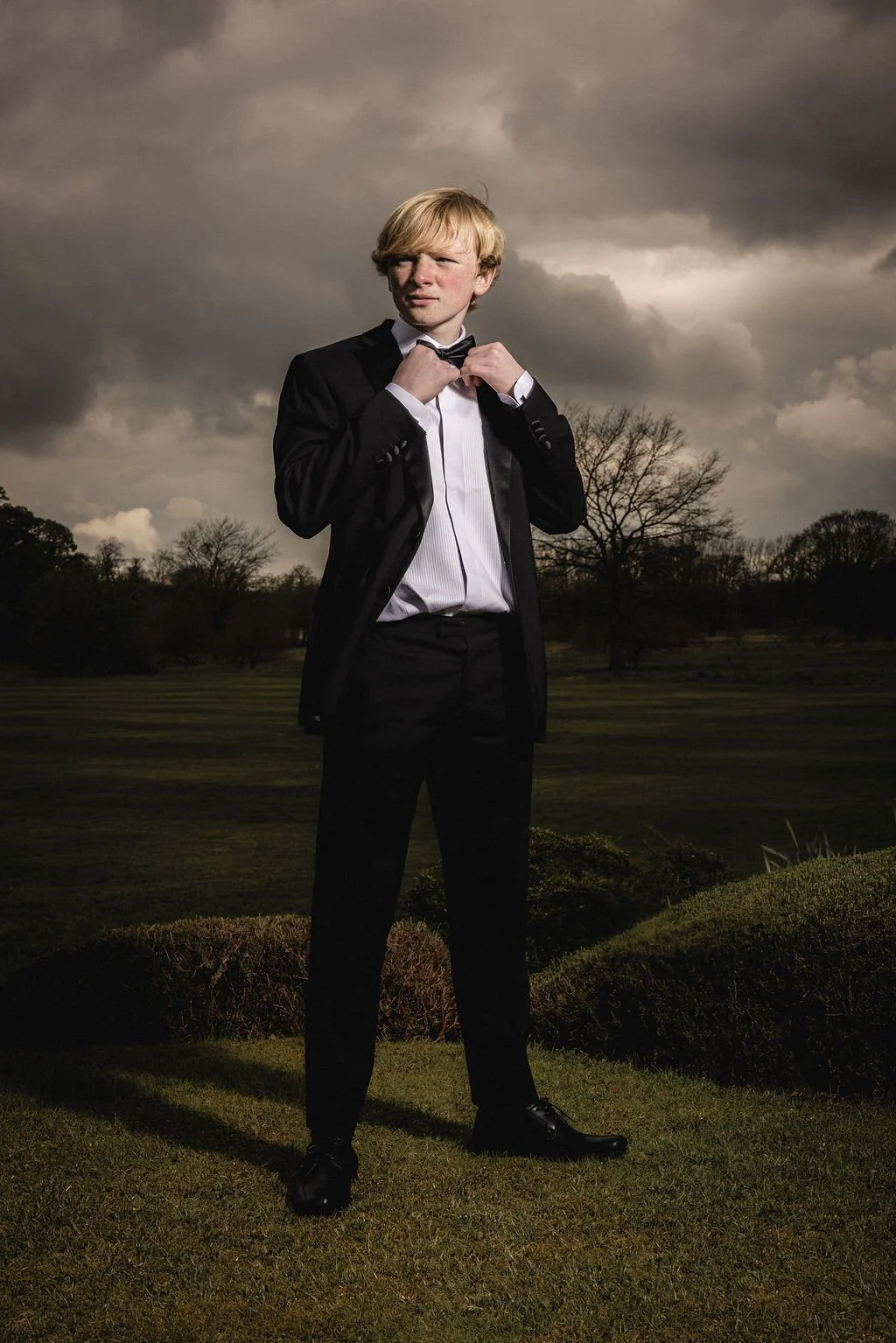 A young man in a tuxedo adjusting his bow tie standing outdoors on a grassy area with trees and dark, cloudy sky in the background.
