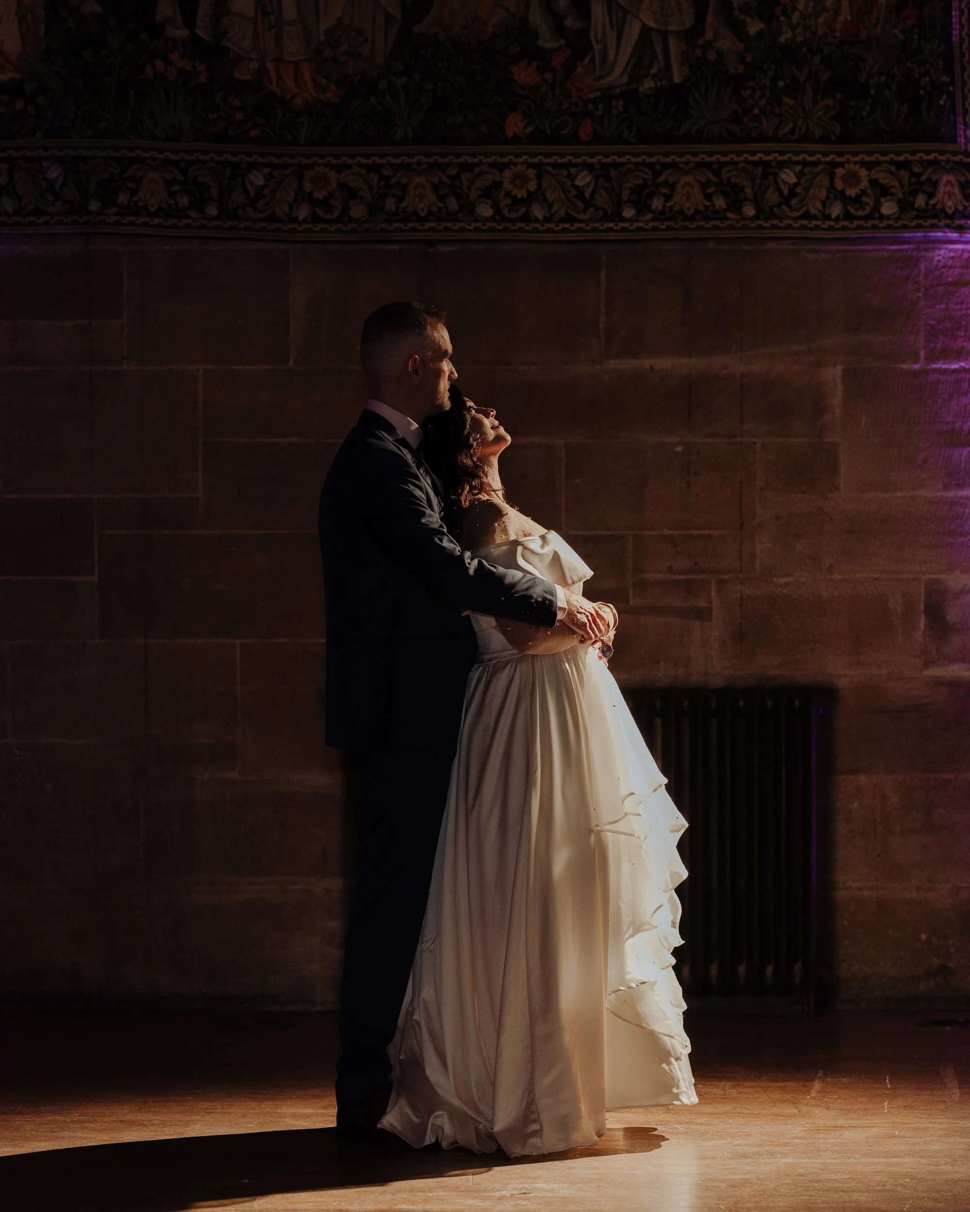 A bride and groom dancing closely together in a dimly lit indoor setting, with the bride in a white gown and the groom in a black suit, illuminated by soft purple and yellow lighting.