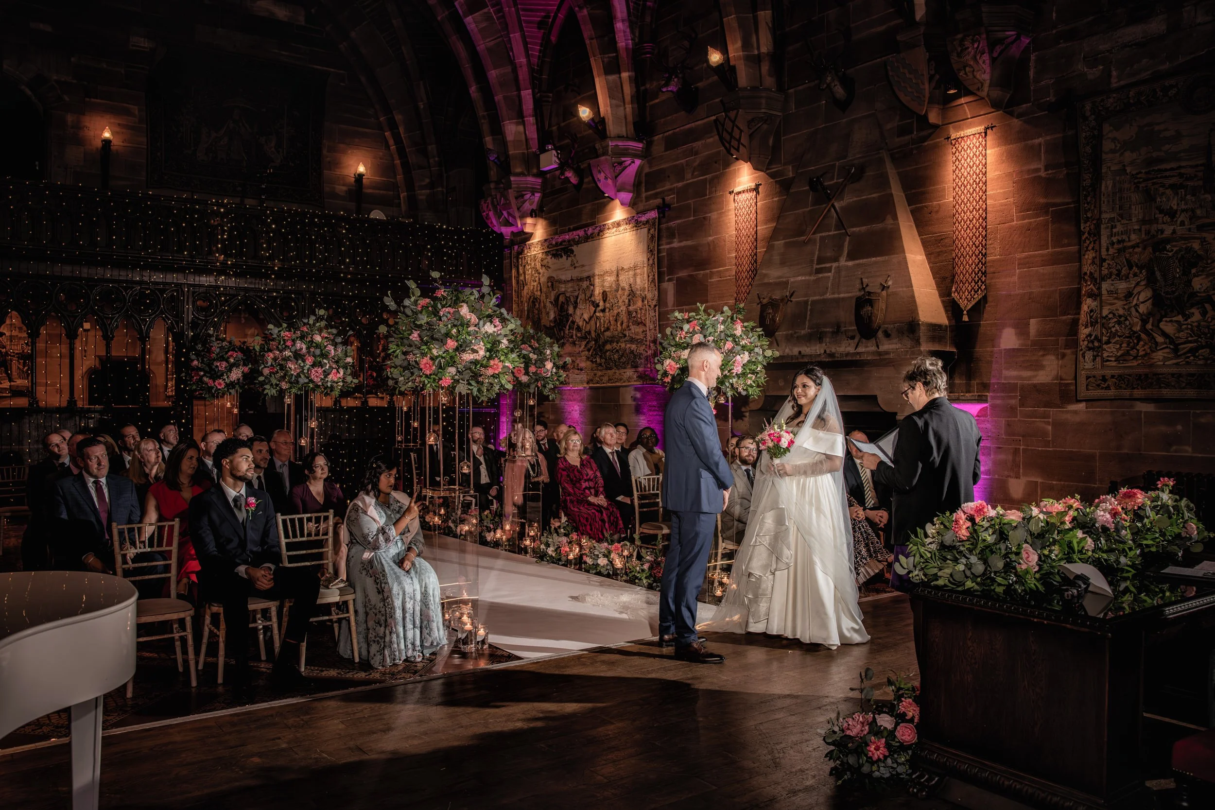 A wedding ceremony taking place inside a decorated hall with the bride and groom standing at the altar, facing each other, with an officiant beside them. Guests are seated on both sides of the aisle, adorned with flowers and lights, with some capturi