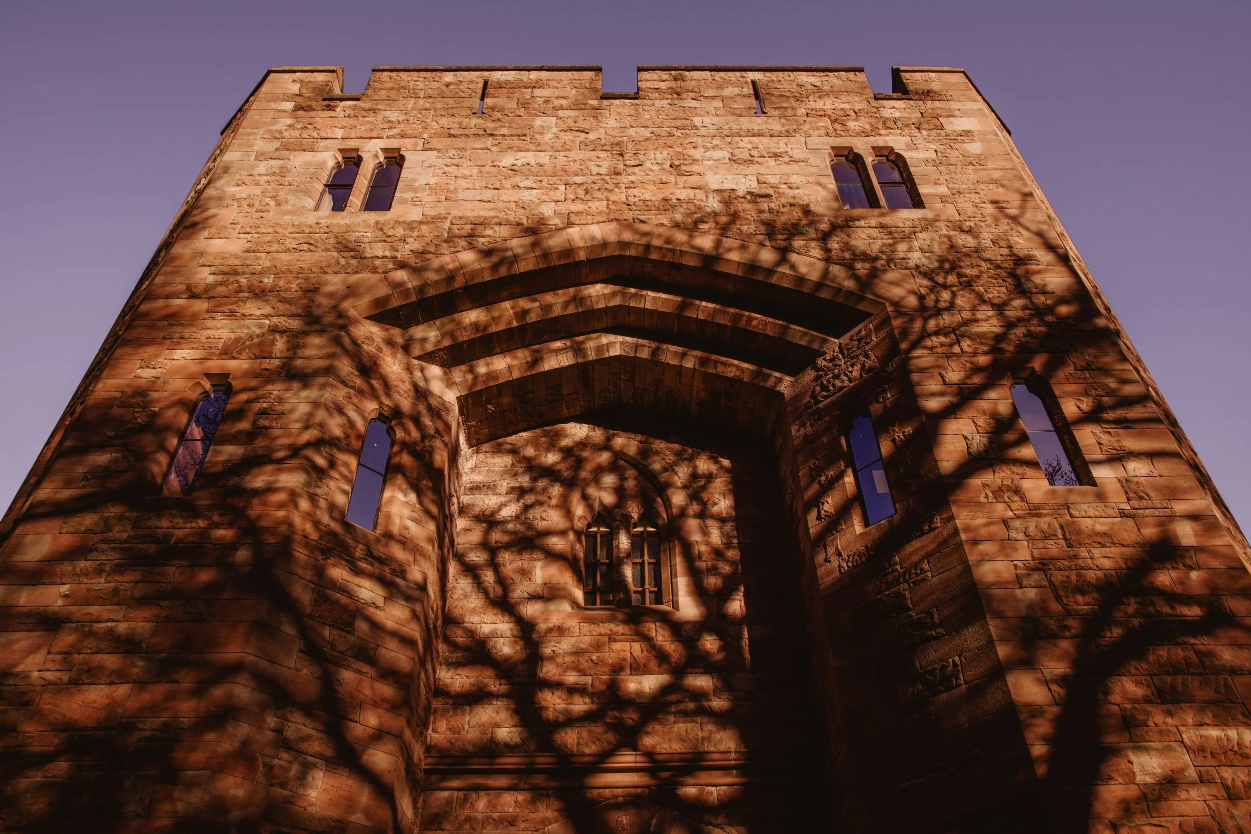 A stone castle with narrow windows at sunset, casting shadows of tree branches on the wall.