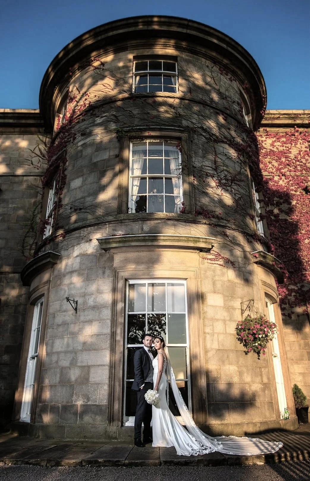 A bride and groom wedding photo standing in front of a large, historic stone building with multiple windows and decorated with hanging flower baskets. The bride wears a white gown with a long train and veil, holding a bouquet of white roses. The groo