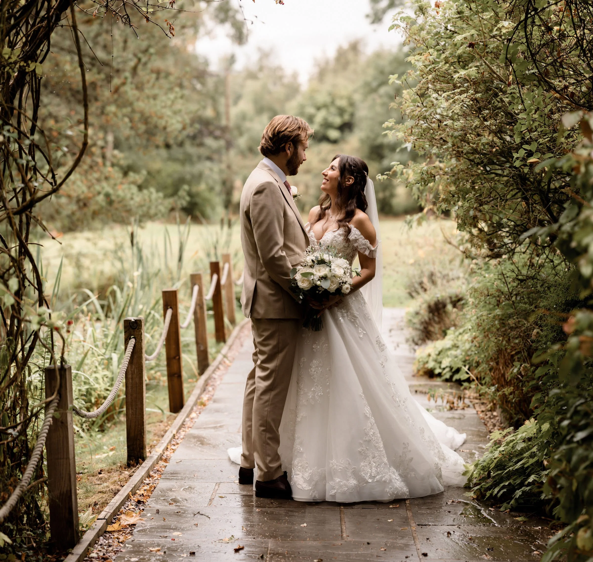 A bride and groom standing on a wooden pathway surrounded by greenery and trees, gazing at each other with smiles.