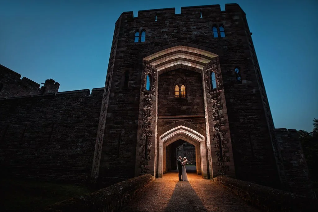 A couple standing under the arch of a large historic stone castle gate during twilight, with a soft glow from an interior window highlighting their embrace.