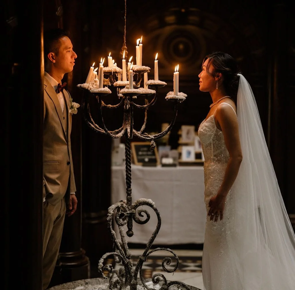 A bride and groom facing each other in a dimly lit room decorated for a wedding, with a large candle chandelier between them.