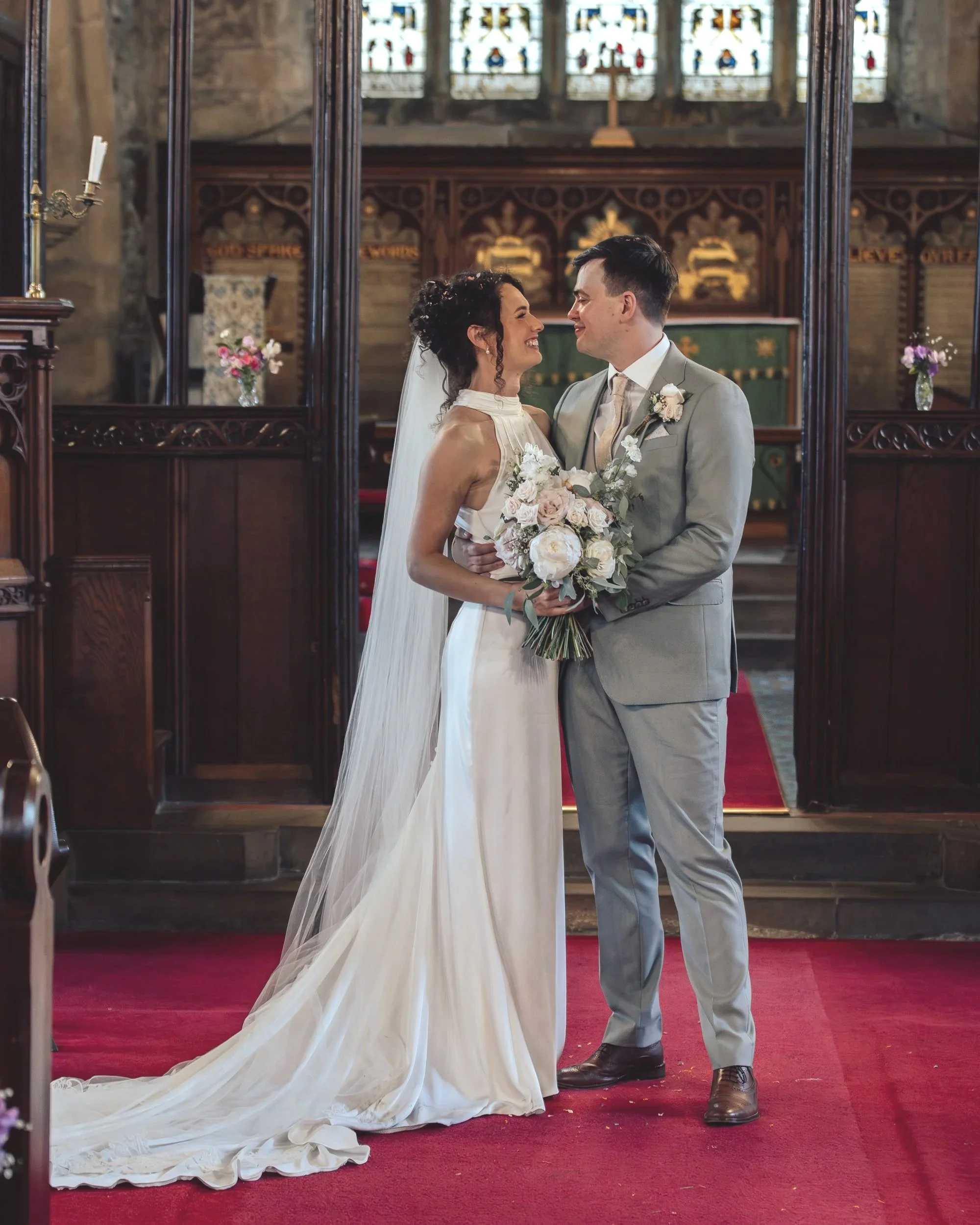 Bride and groom smiling at each other in a church, standing on a red carpet with wooden pews and stained glass windows in the background.