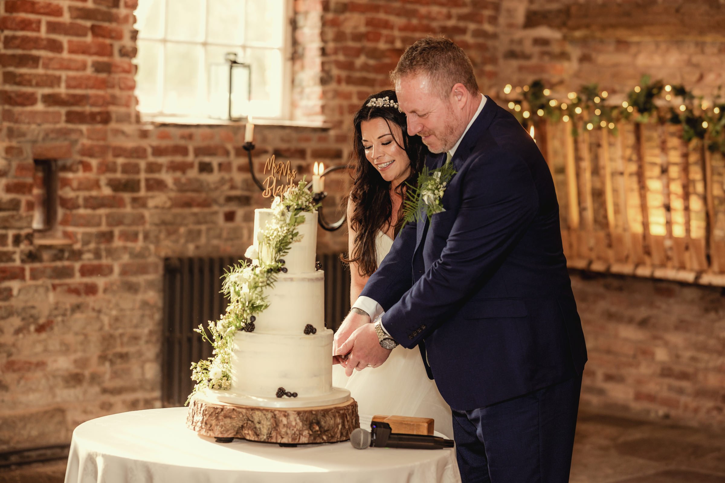 A newlywed couple cuts their wedding cake together, smiling, in a rustic indoor setting with a brick wall background and wedding decorations.