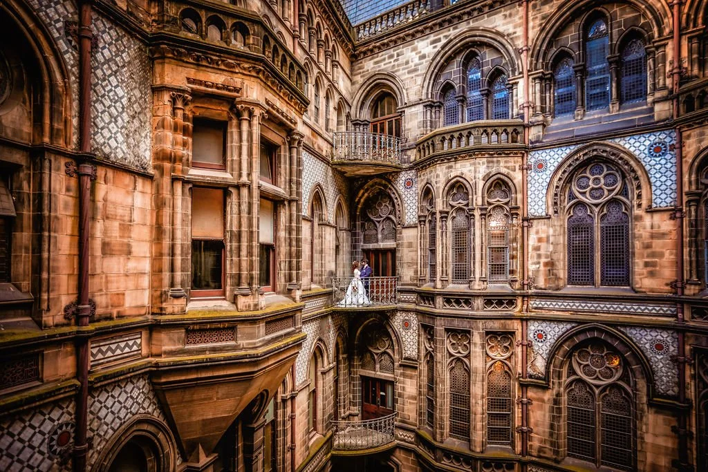 A bride and groom standing on a small balcony inside a historic, ornate Gothic-style cathedral with stained glass windows and intricate stonework.