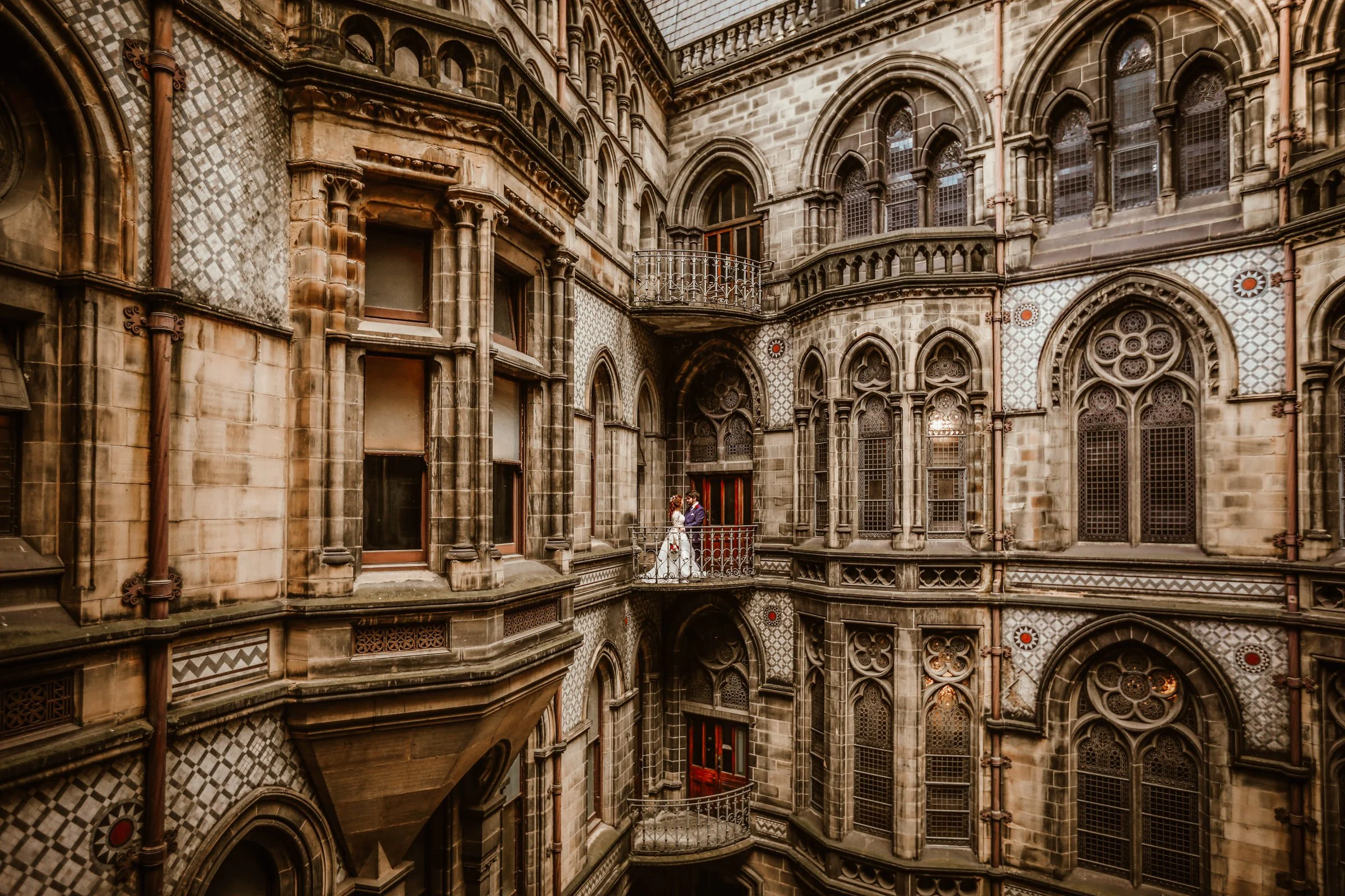 Couple in wedding attire standing on a small balcony inside a historic Gothic-style cathedral with intricate stained glass windows and ornate stonework.