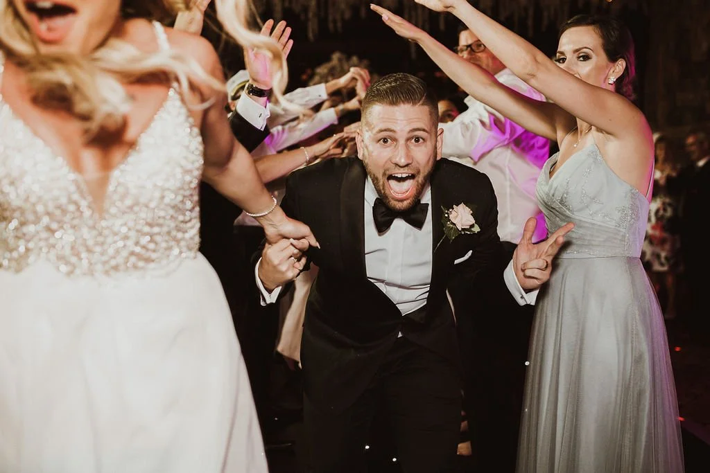 People dancing at a wedding reception, with a man in a tuxedo in the center making a joyful expression and holding hands with women in formal dresses.
