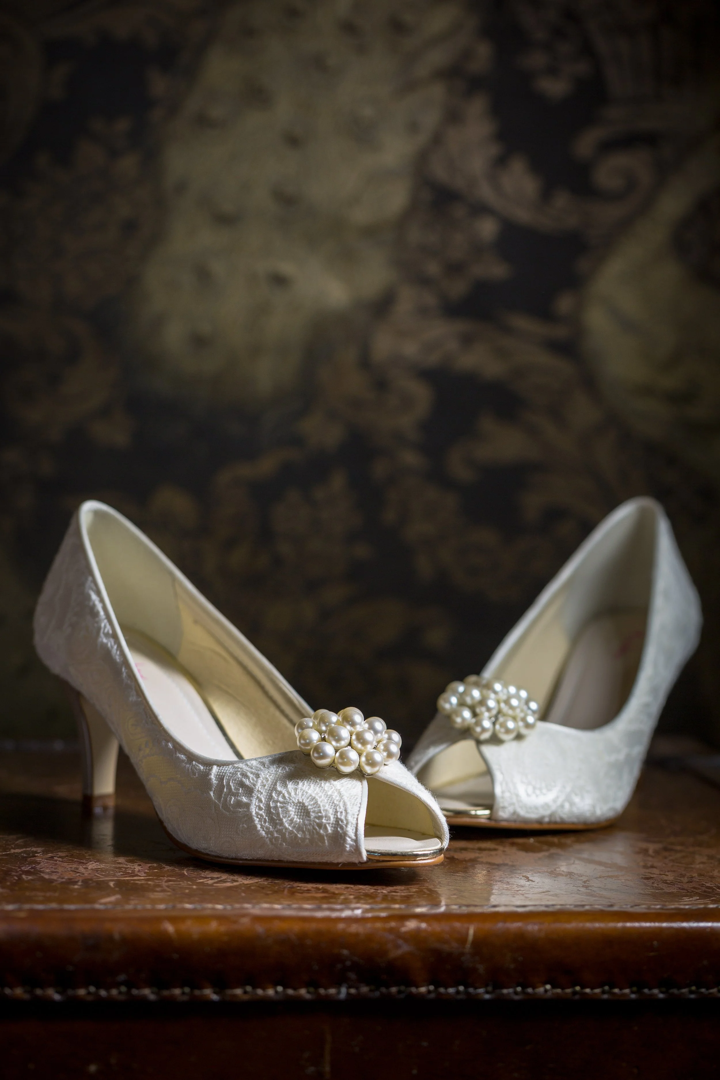 White lace wedding shoes on an old brown chest against a peacock patterned luxury wallpaper
