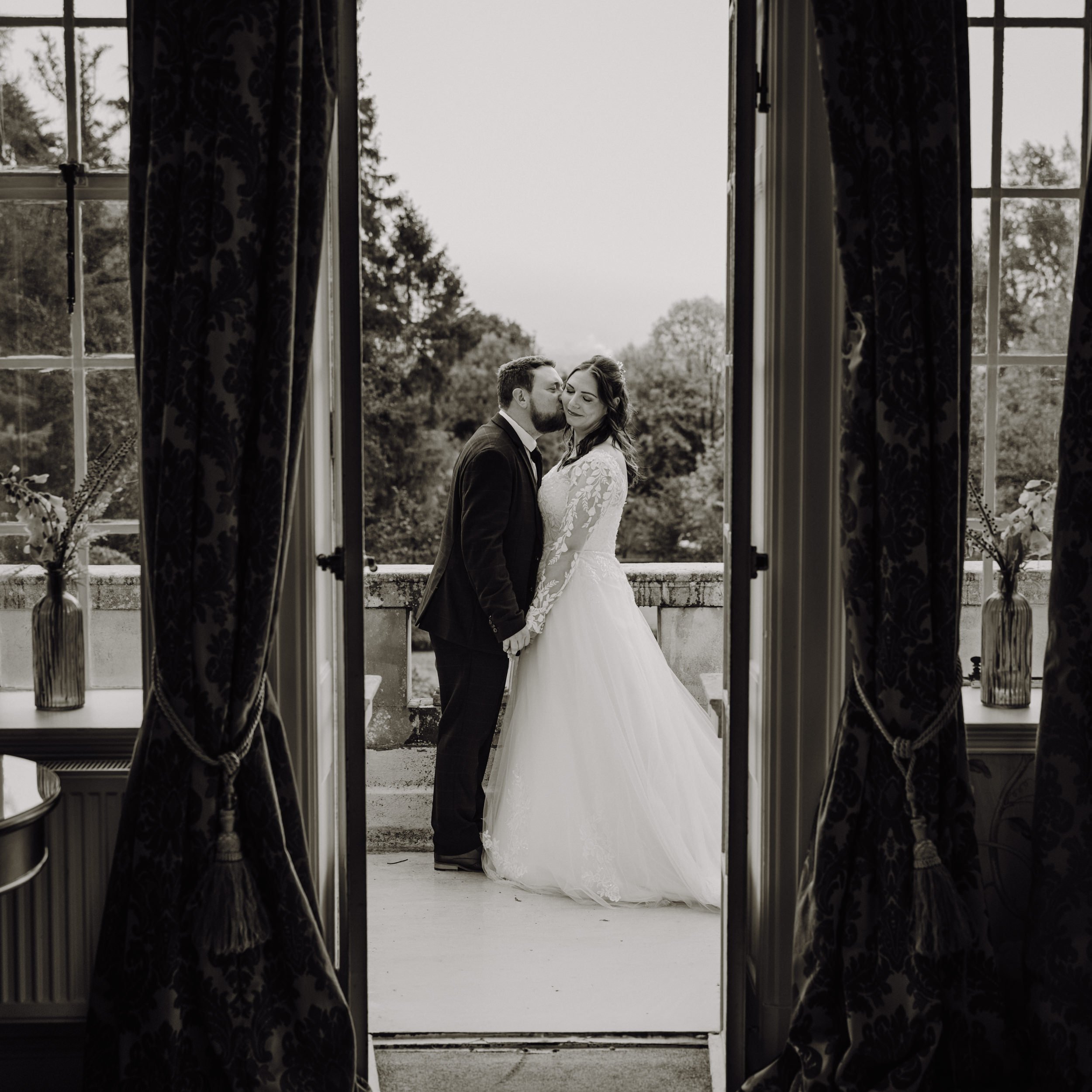 A black-and-white photo of a bride and groom standing outside on a balcony, holding hands and sharing a kiss, framed by open curtains inside a room with large windows.