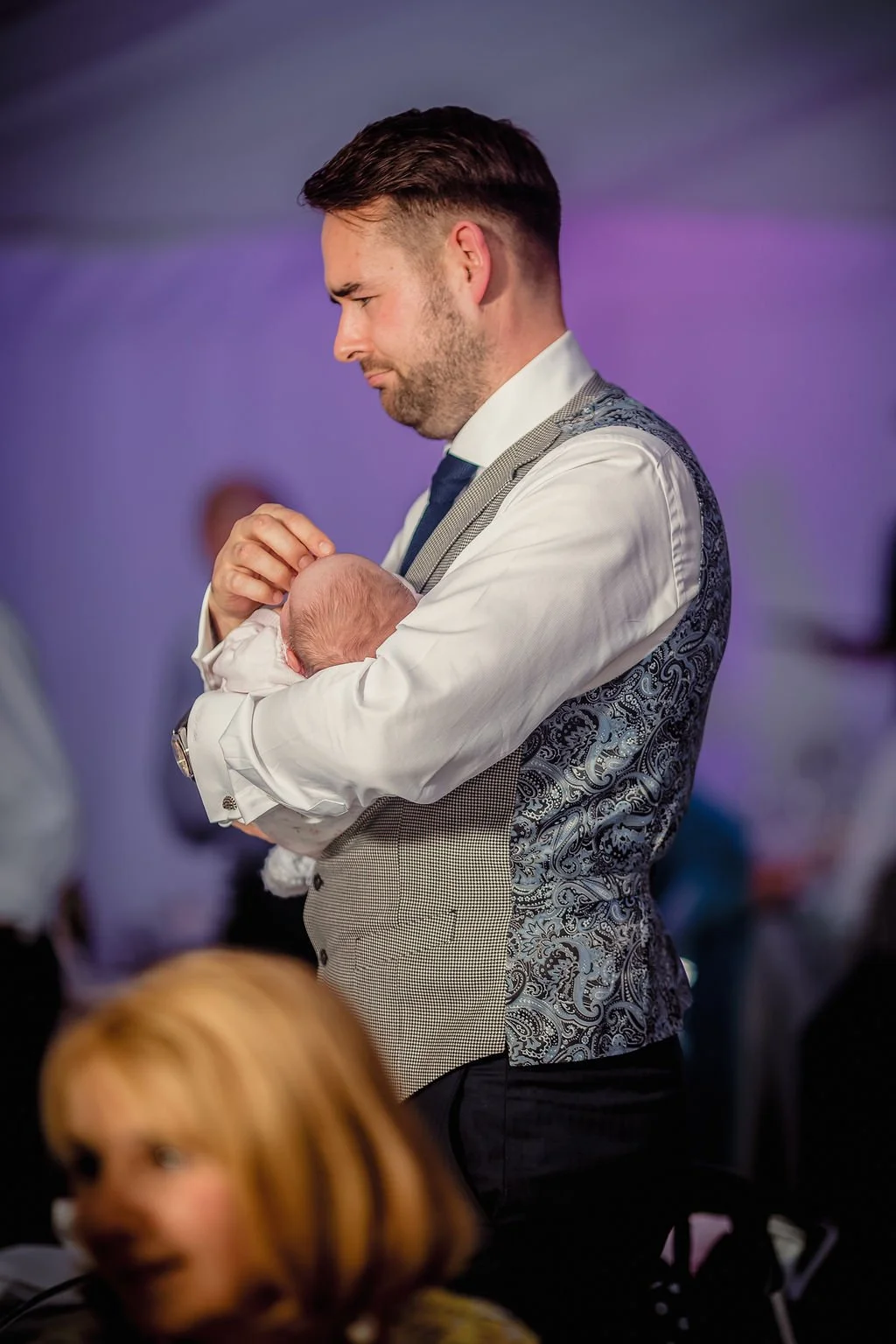 A man holding a newborn baby in his arms at an event with dim lighting and purple hues in the background.