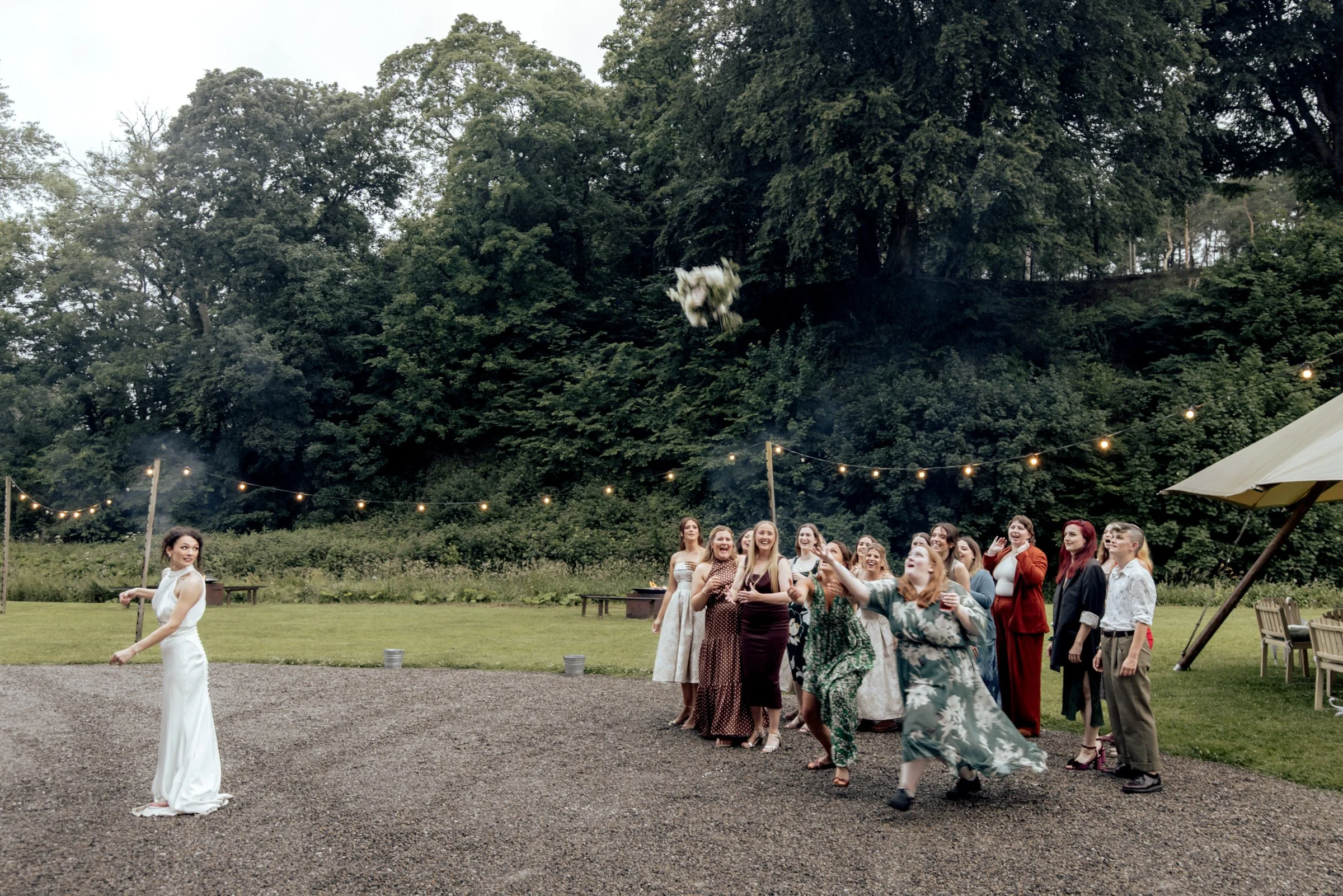 A group of women celebrating outdoors, some standing and some in a line, with a bride in a white wedding dress in the foreground, during a wedding or party at a scenic venue with trees, string lights, and a tent.