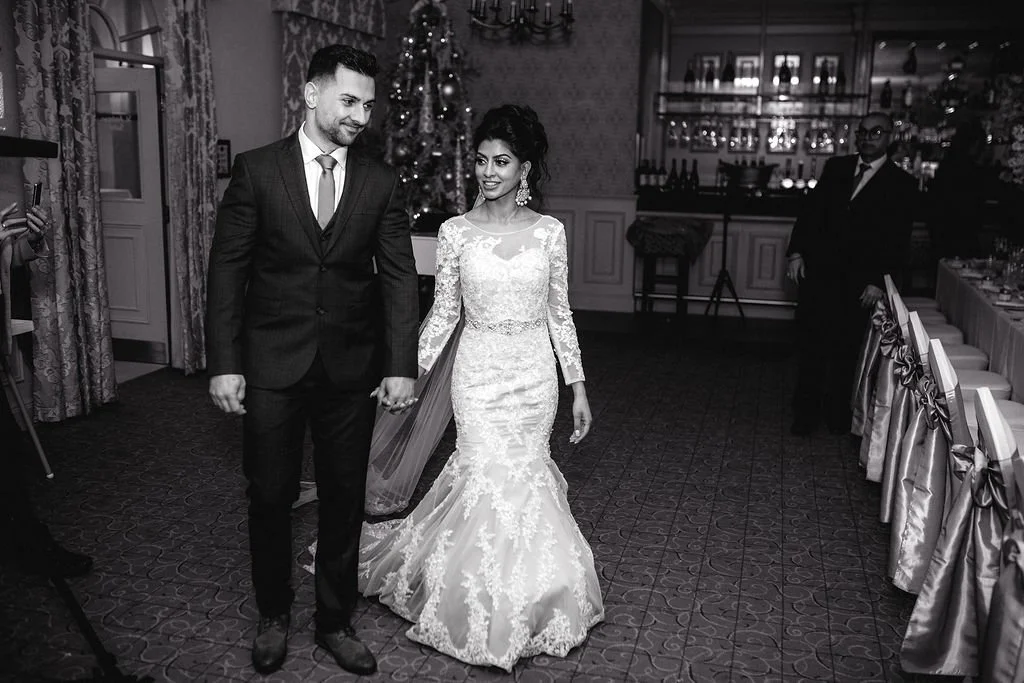 A bride and groom walking hand-in-hand at their wedding reception in a decorated banquet hall, with a Christmas tree in the background.