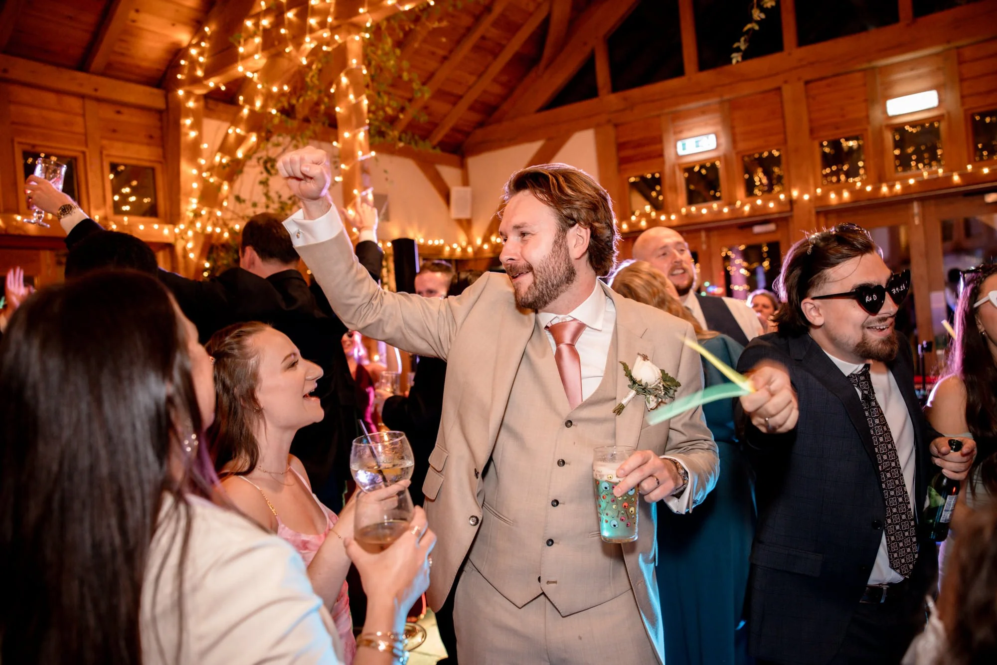 People celebrating at a wedding reception in a wooden hall decorated with string lights, with a man in a beige suit and tie holding a drink and raising his fist in celebration, surrounded by smiling guests.