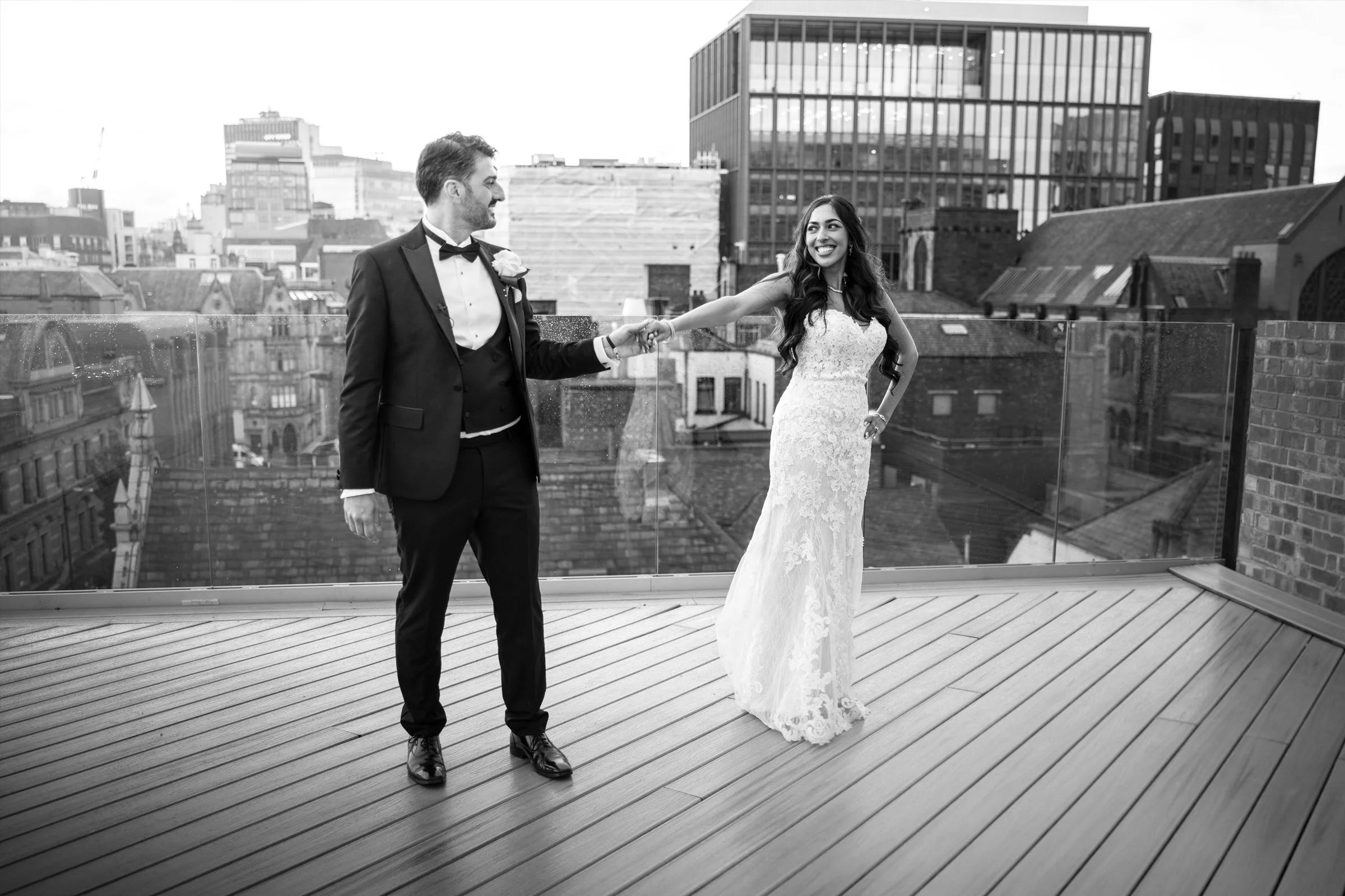 A newlywed couple on a rooftop terrace, holding hands and smiling at each other, with city buildings in the background.