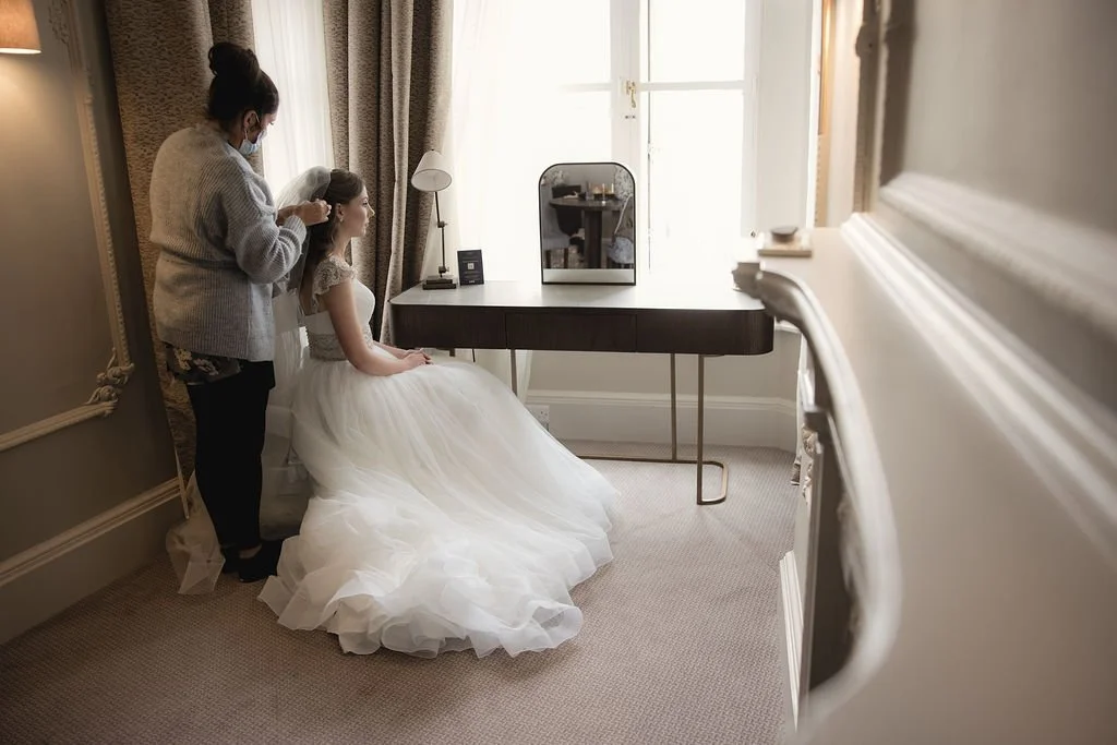 A bride in a white wedding dress sitting on a chair while a woman styles her hair in a bedroom with natural light, a mirror, and elegant decor.