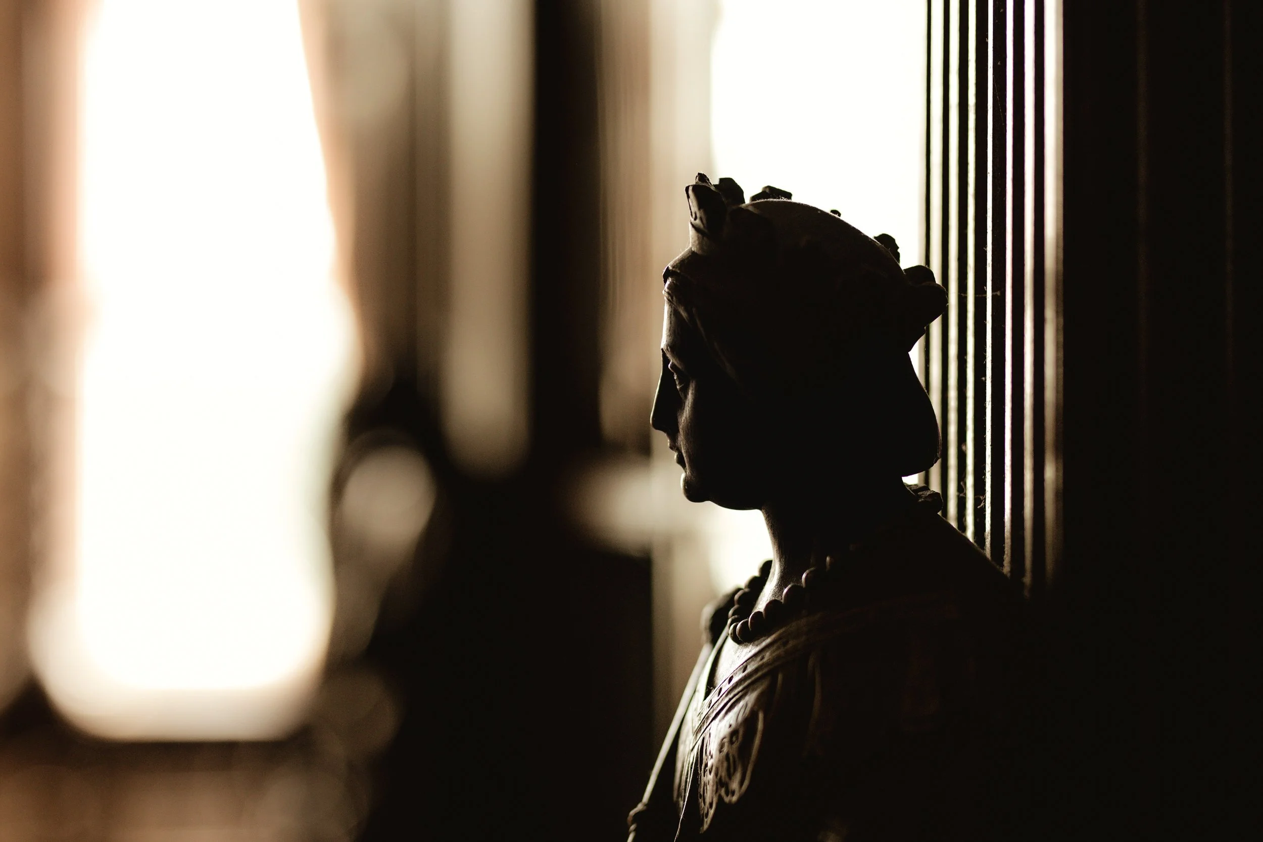 Silhouette of a woman wearing a head wrap and necklace, sitting against a striped wall near a window with bright light, creating a shadowed profile.