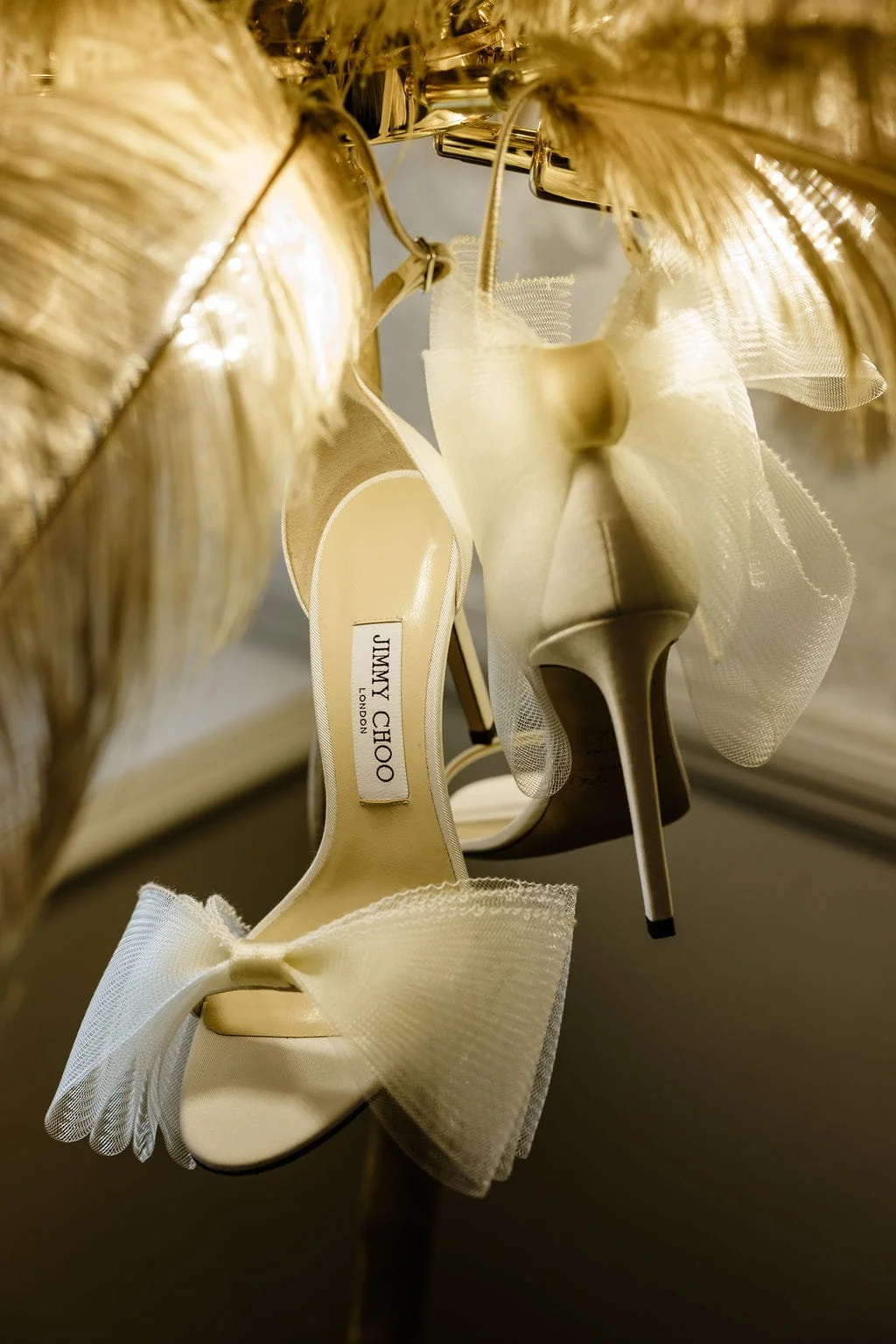 A pair of white Jimmy Choo high-heeled shoes with bows, hanging upside down, viewed from below, with a gold decorative item in the background.