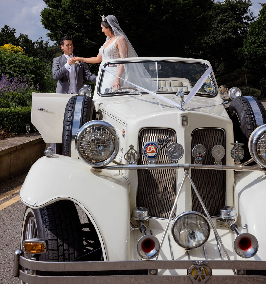 Bride and groom wearing wedding attire exchanging rings on a vintage white car decorated with ribbons, outdoors with trees and flowers in the background.