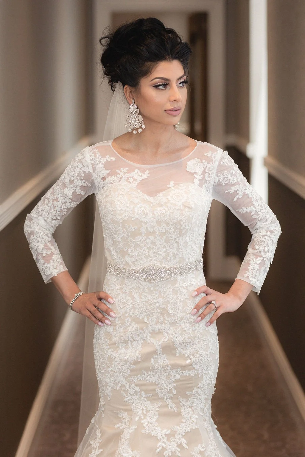 A woman in a white lace wedding dress standing in a hallway with hands on her hips.