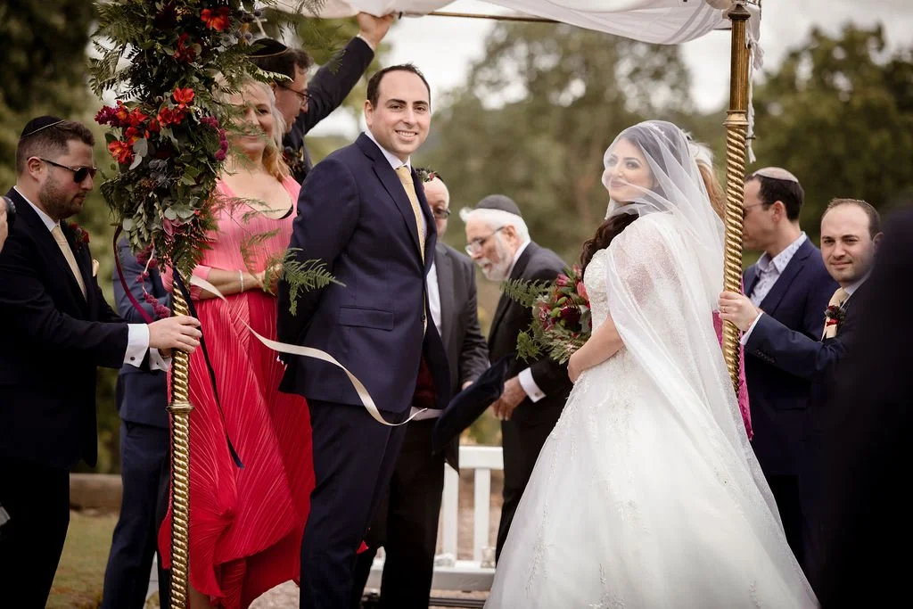 A wedding scene outdoors with a bride and groom under a decorated canopy, surrounded by wedding party members, mostly men in suits and women in dresses, with some holding ribbons in a ceremonial setting.
