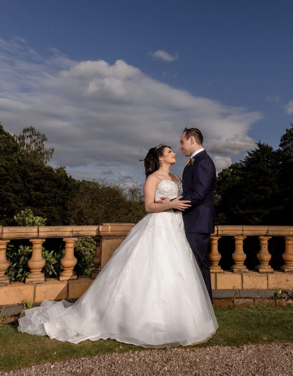 A bride and groom stand close together outdoors on a stone patio, gazing into each other's eyes, with trees and a cloudy sky in the background.