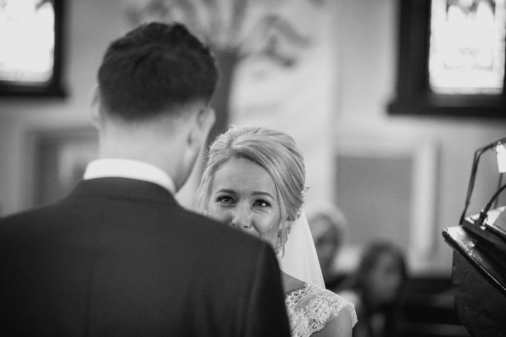 A bride smiling at a groom during a wedding ceremony, with the bride's face visible and the groom's back facing the camera.