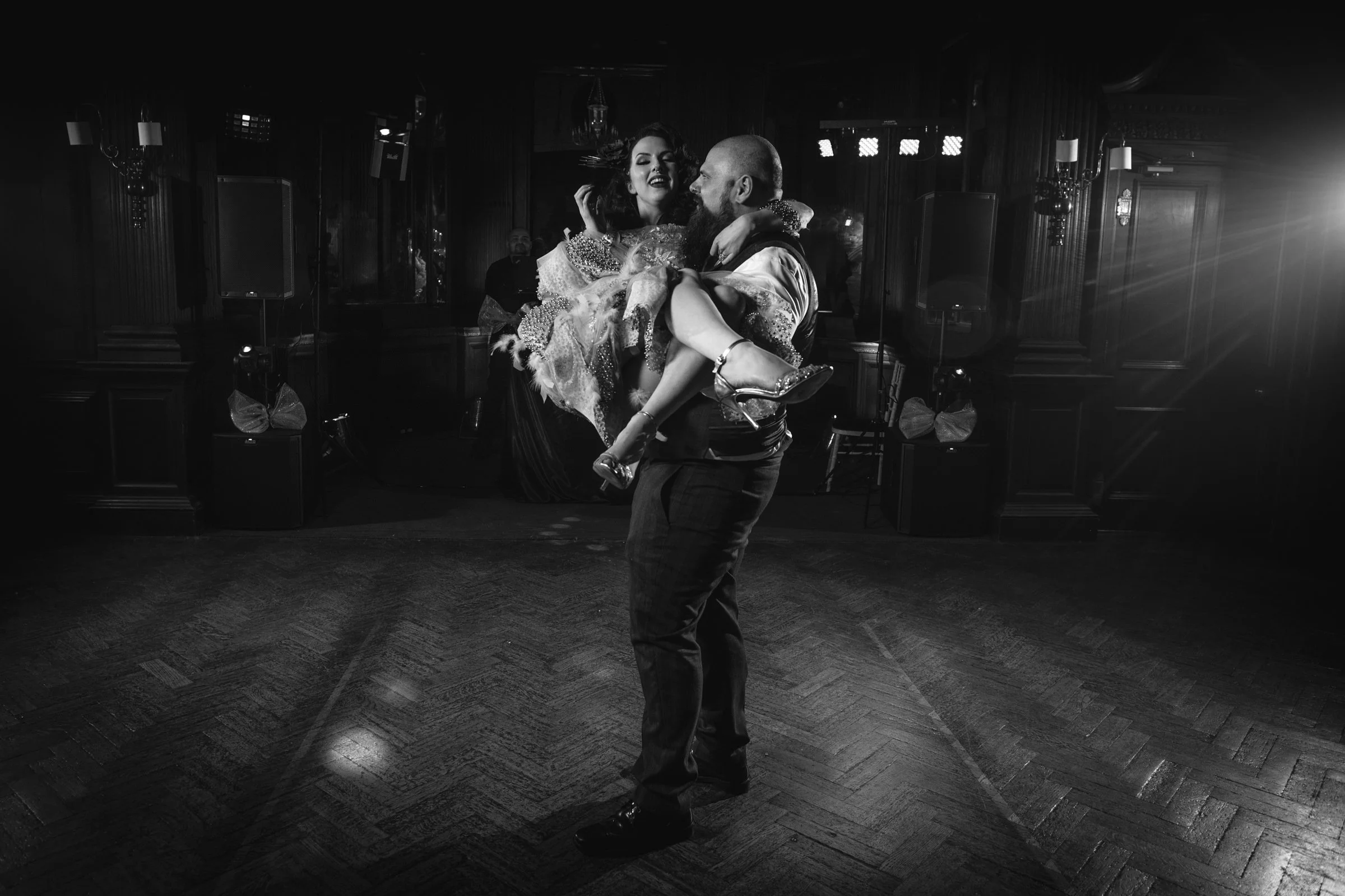 A man is dancing with a woman in a fancy dress on a dance floor in a dimly lit room with wooden paneling and DJ equipment in the background.