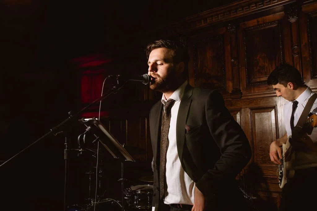 A man in a black suit with a white shirt singing into a microphone on a stand, with another man in a white shirt and tie playing an electric guitar in the background, in a wood-paneled room.