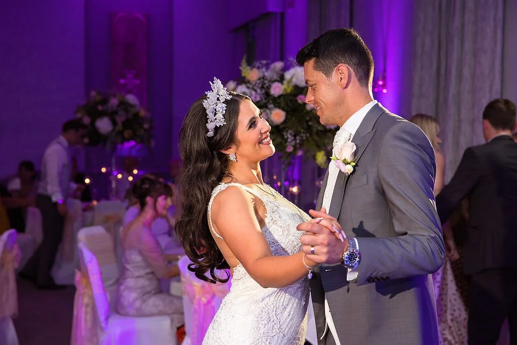 A bride and groom dancing at their wedding reception with purple lighting and floral arrangements in the background