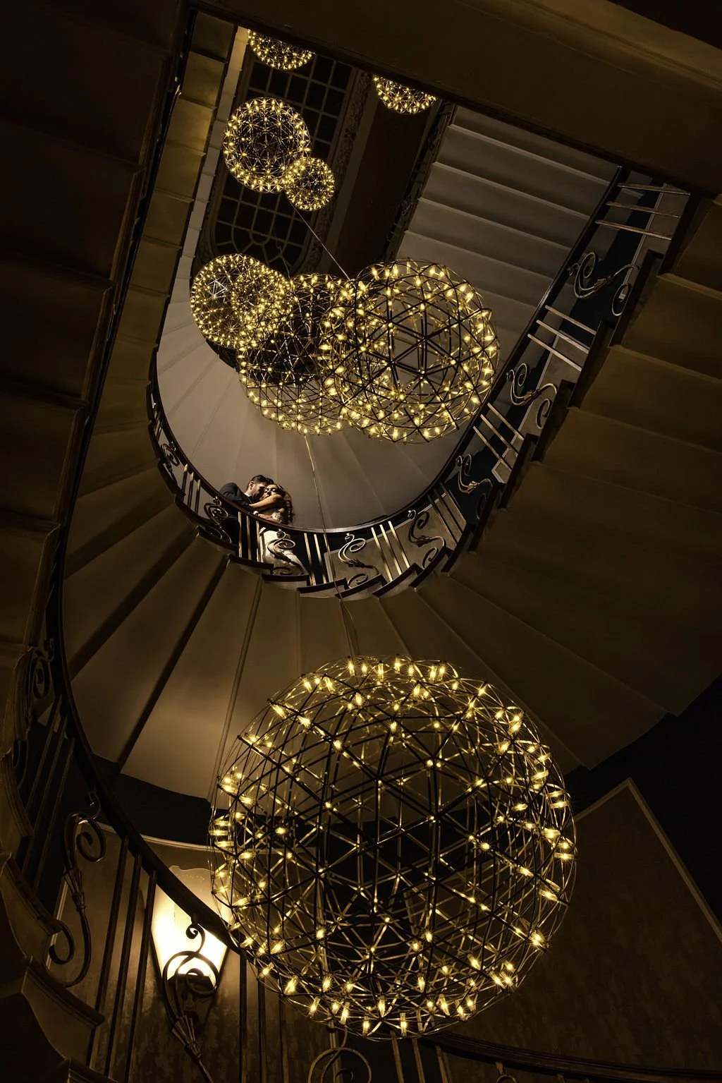 View of a spiral staircase from below, decorated with large spherical pendant lights with string lights inside, creating a geometric pattern in a dimly lit interior.